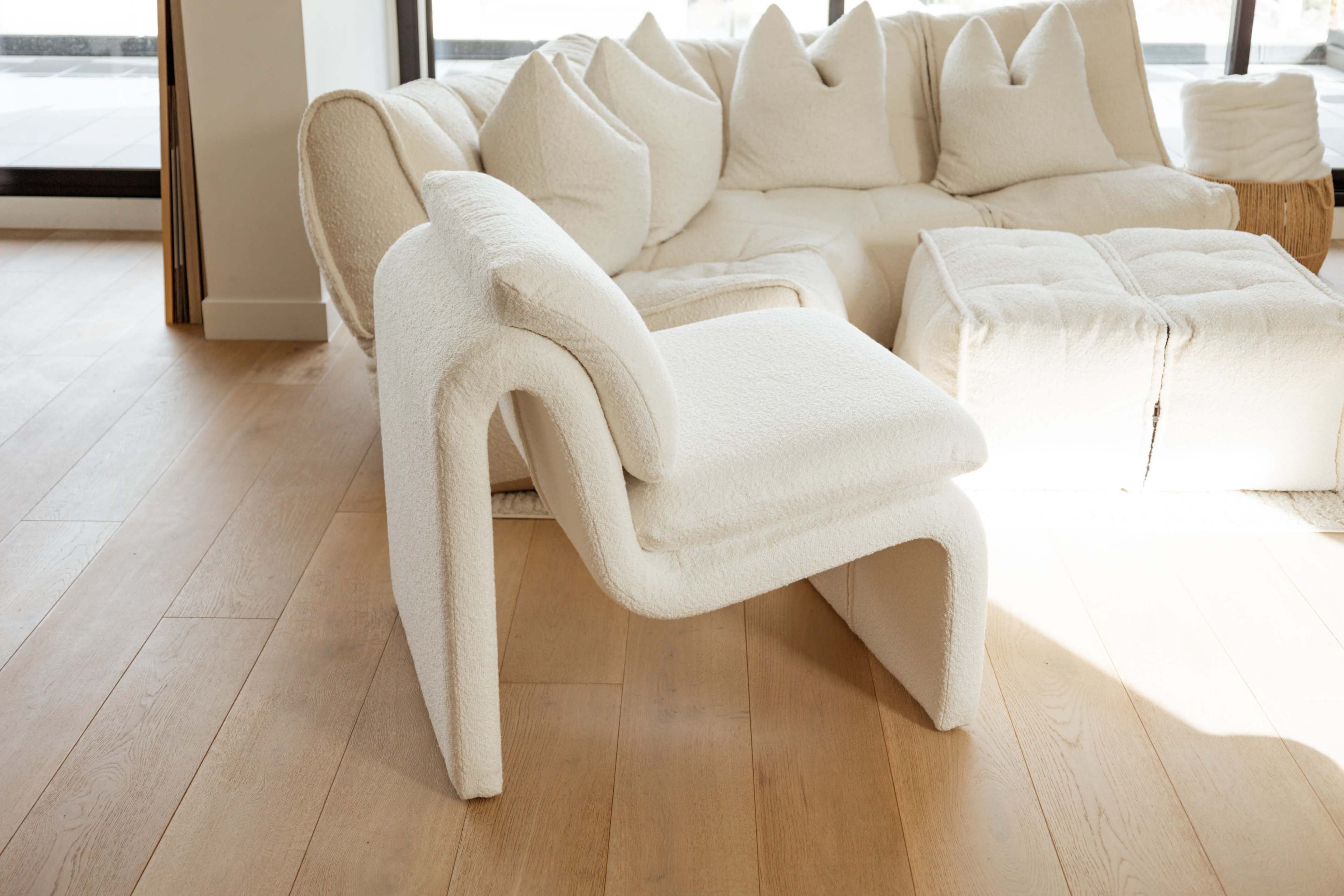 A white, textured chair positioned beside a light-colored sofa in a spacious, sunlit room with wooden flooring.