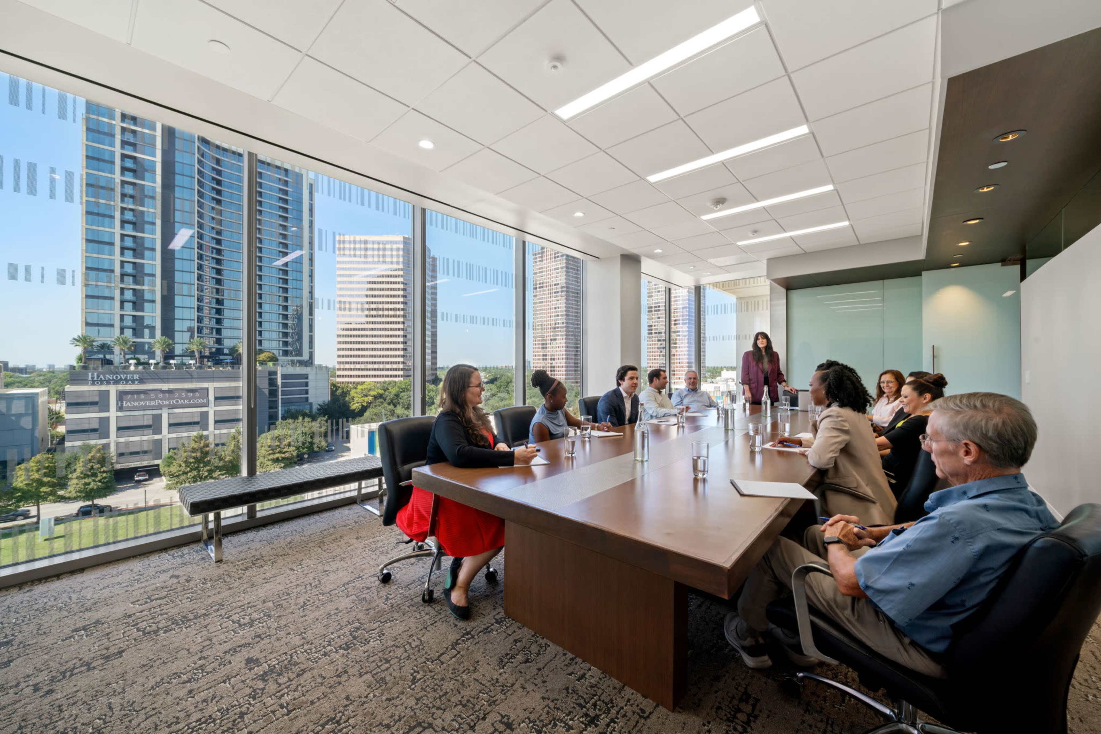 A group of people is seated around a large conference table in a modern office with a view of city skyscrapers through large windows.
