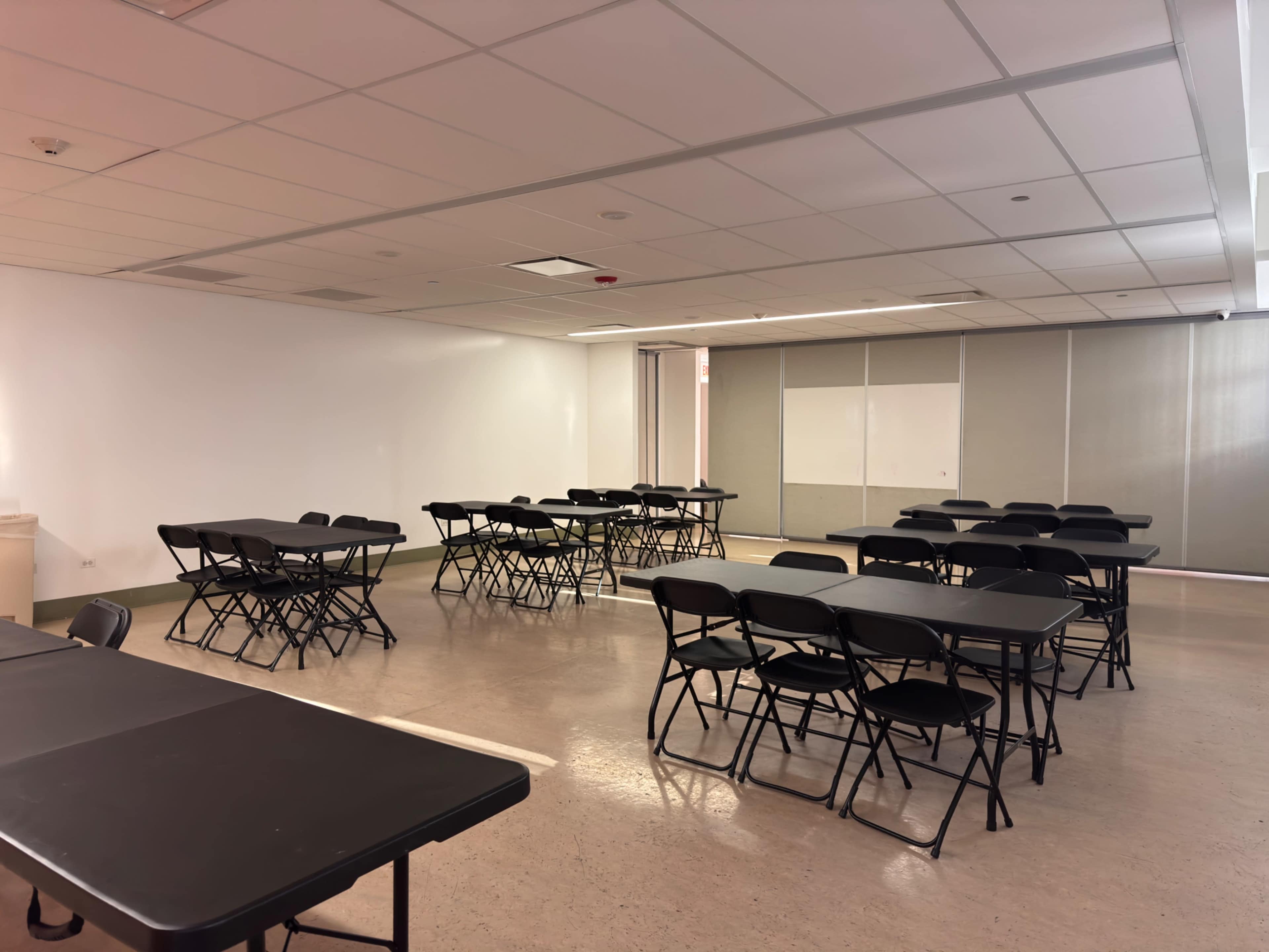 The image shows a large, empty room set up with several black folding tables and chairs arranged in a grid pattern.