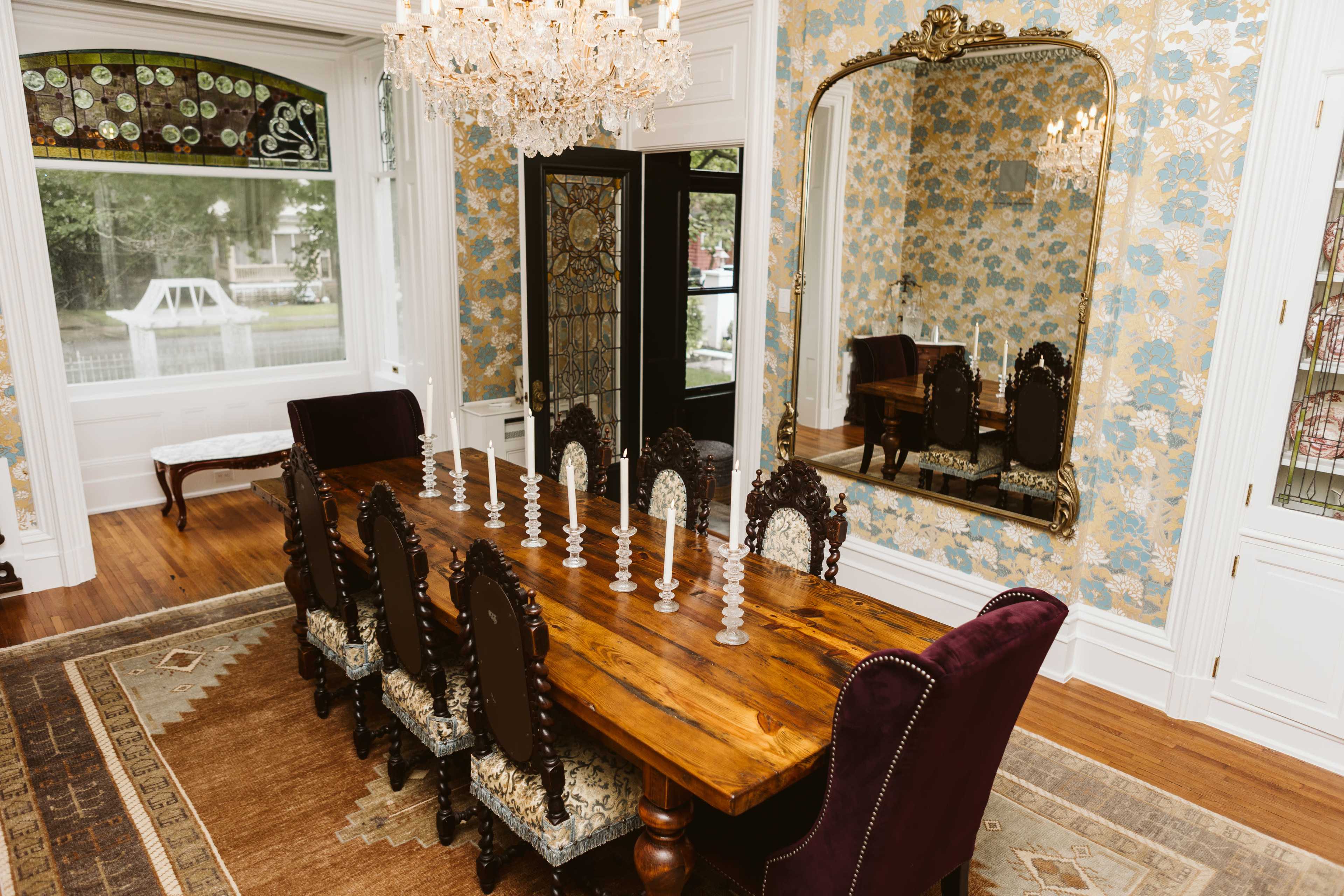 The image depicts a dining room featuring a large wooden table surrounded by ornate chairs, with a crystal chandelier and a large mirror reflecting the room's floral wallpaper.