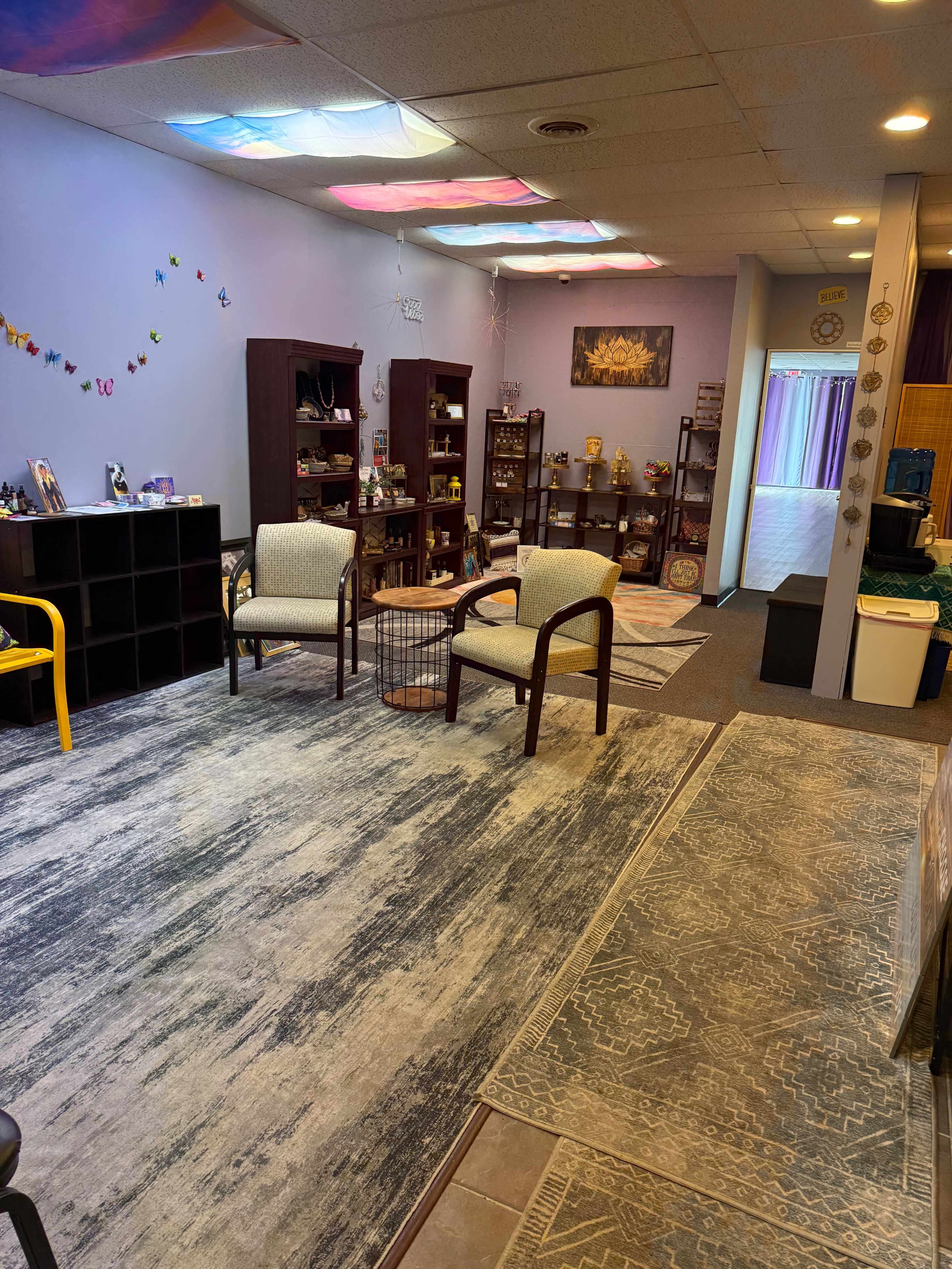 The image shows a softly lit interior space with shelves displaying various items, chairs arranged for seating, and a large area rug on the floor.