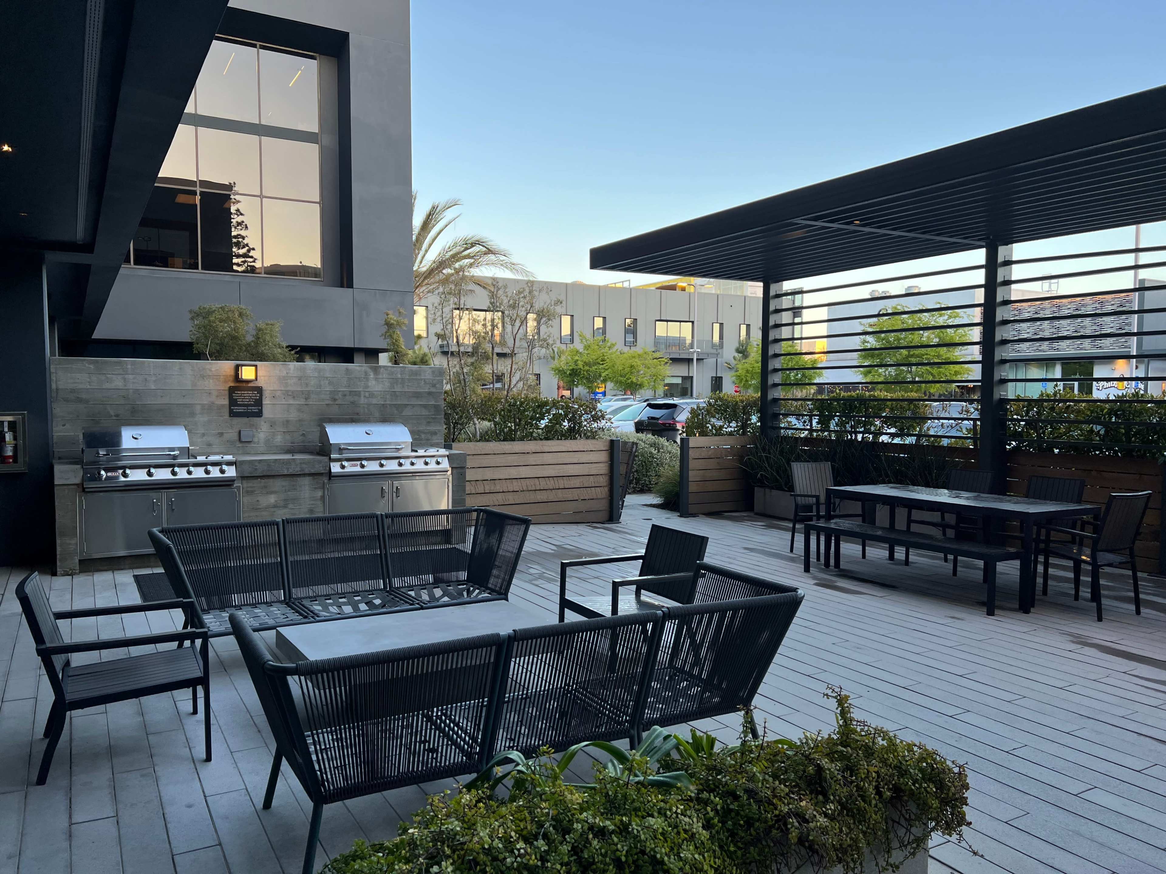 The scene shows a modern outdoor patio area with two barbecue grills, black seating, and tables surrounded by greenery and a building in the background.