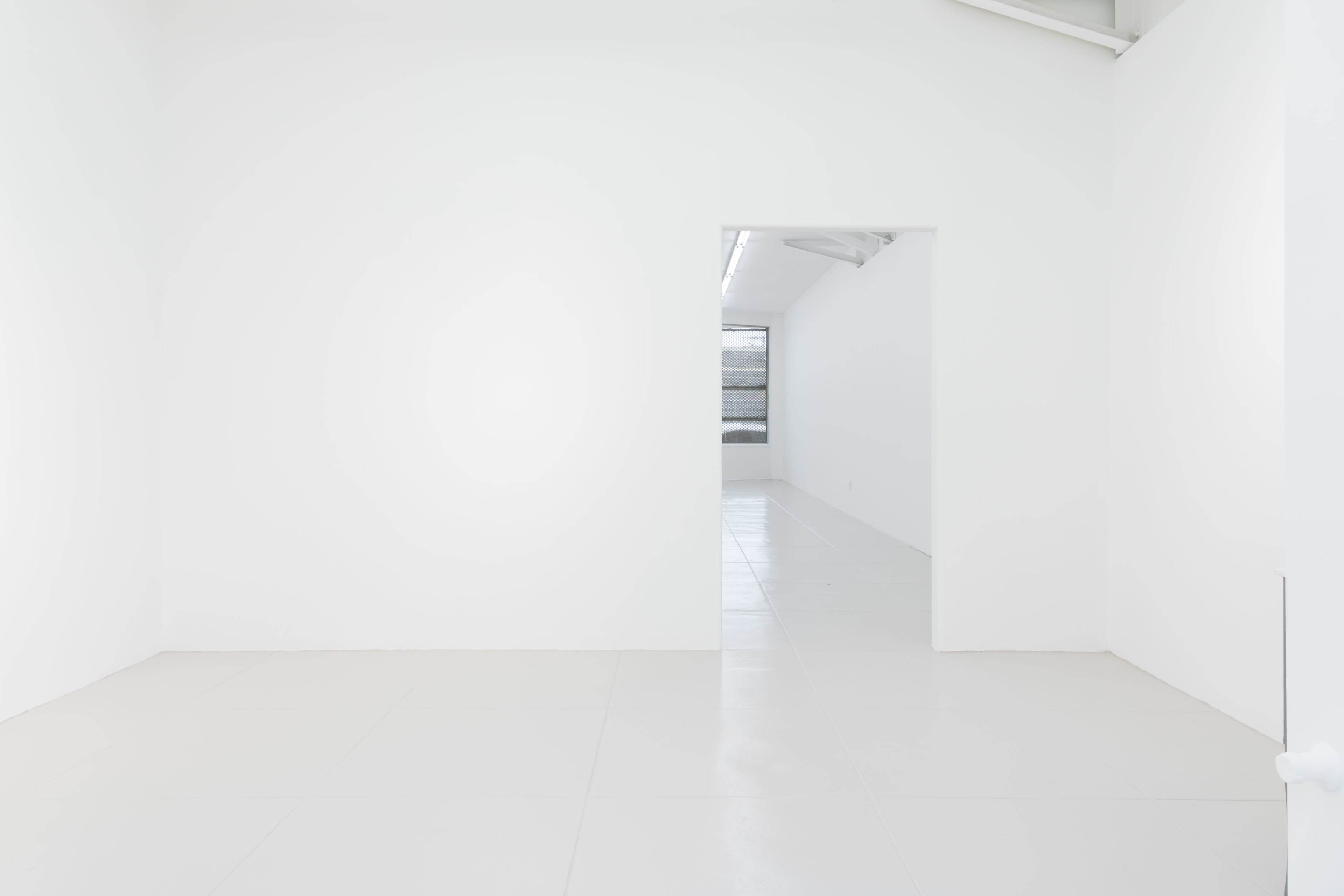 A minimalist, empty room with white walls and a doorway leading to another bright space.