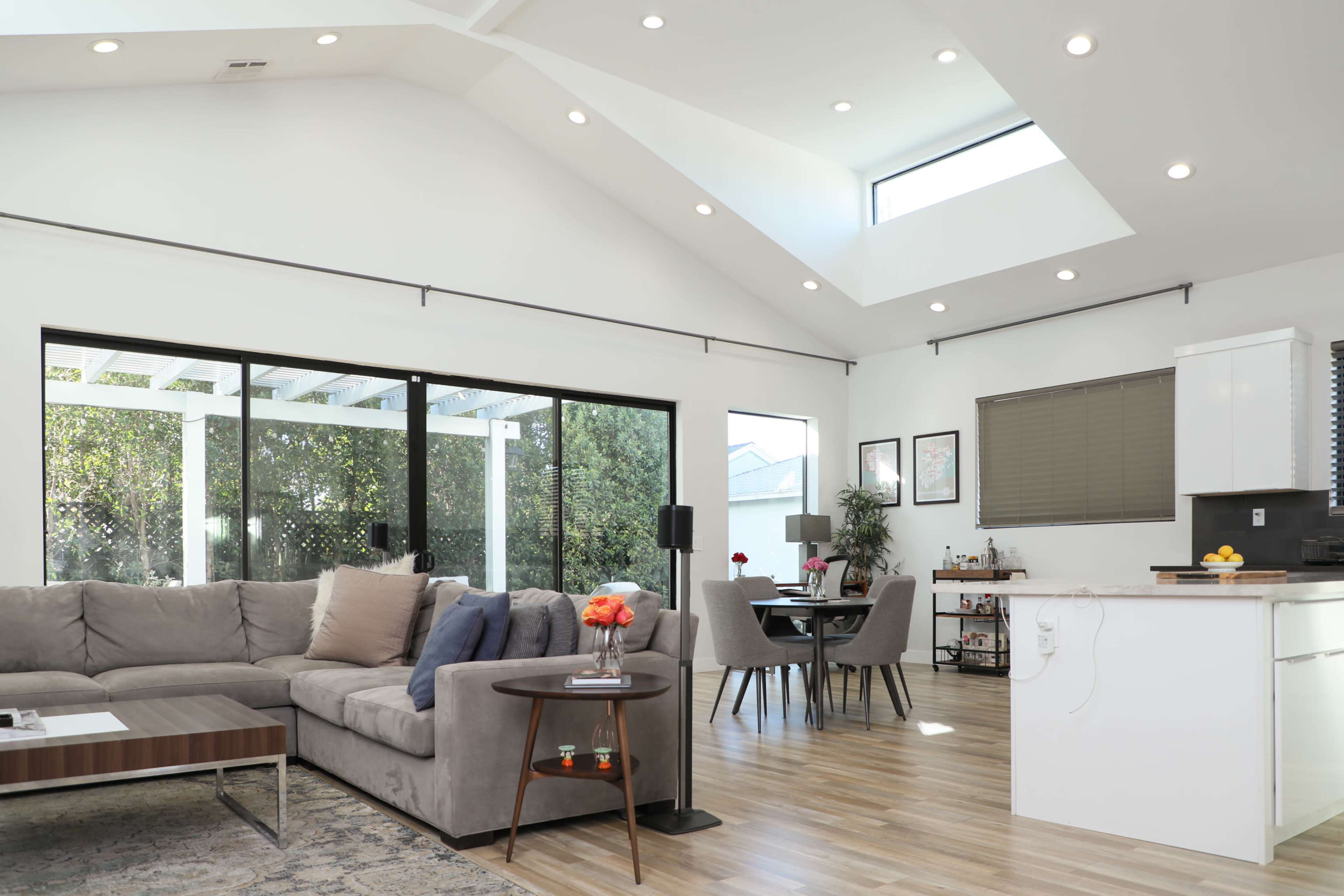 A bright, modern living space featuring a gray sectional sofa, a dining table, and large windows that allow natural light to fill the room.