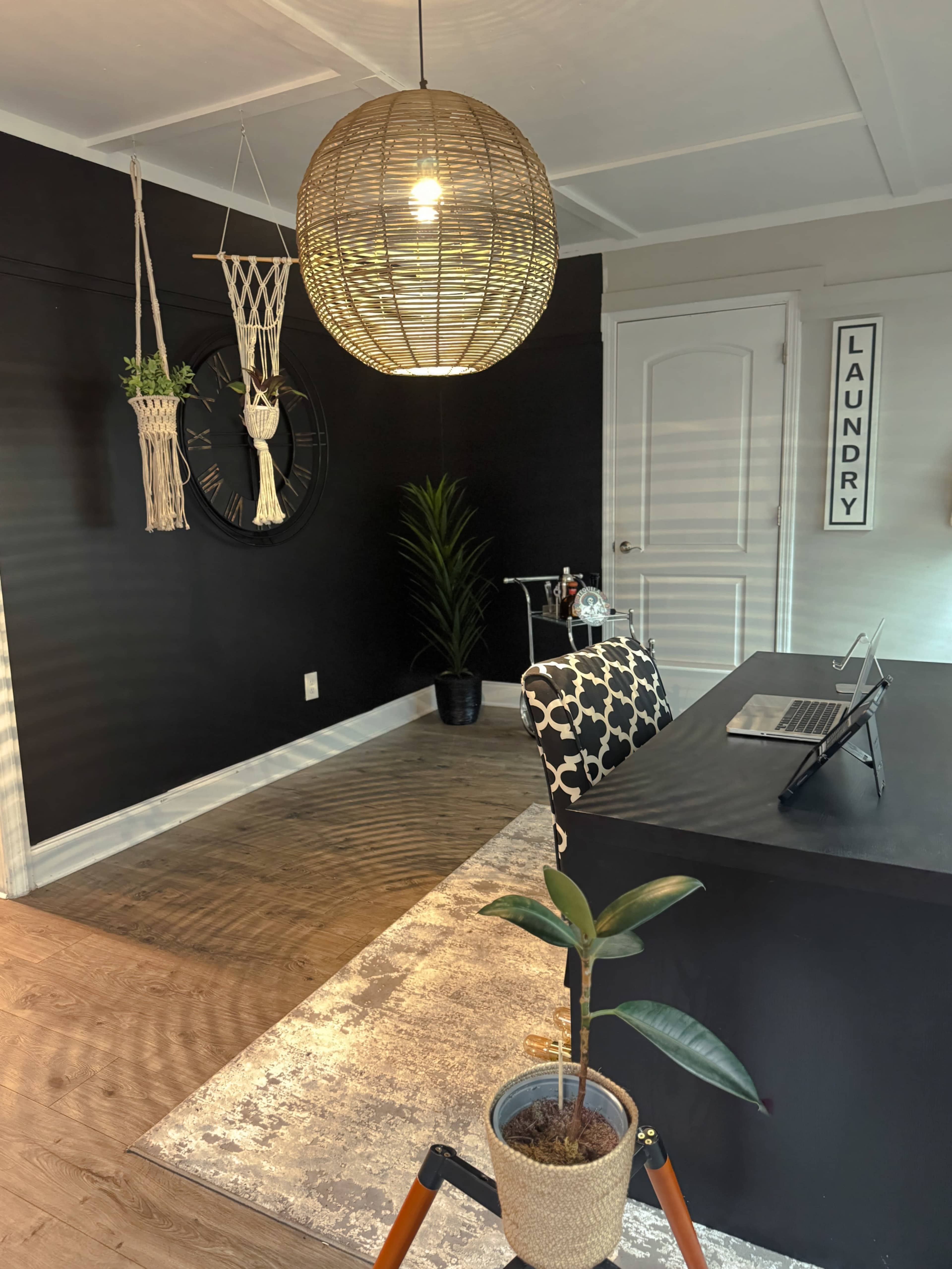 The image shows a modern home office with a large pendant light, a black accent wall, and a desk with a laptop, surrounded by plants and decorative elements.