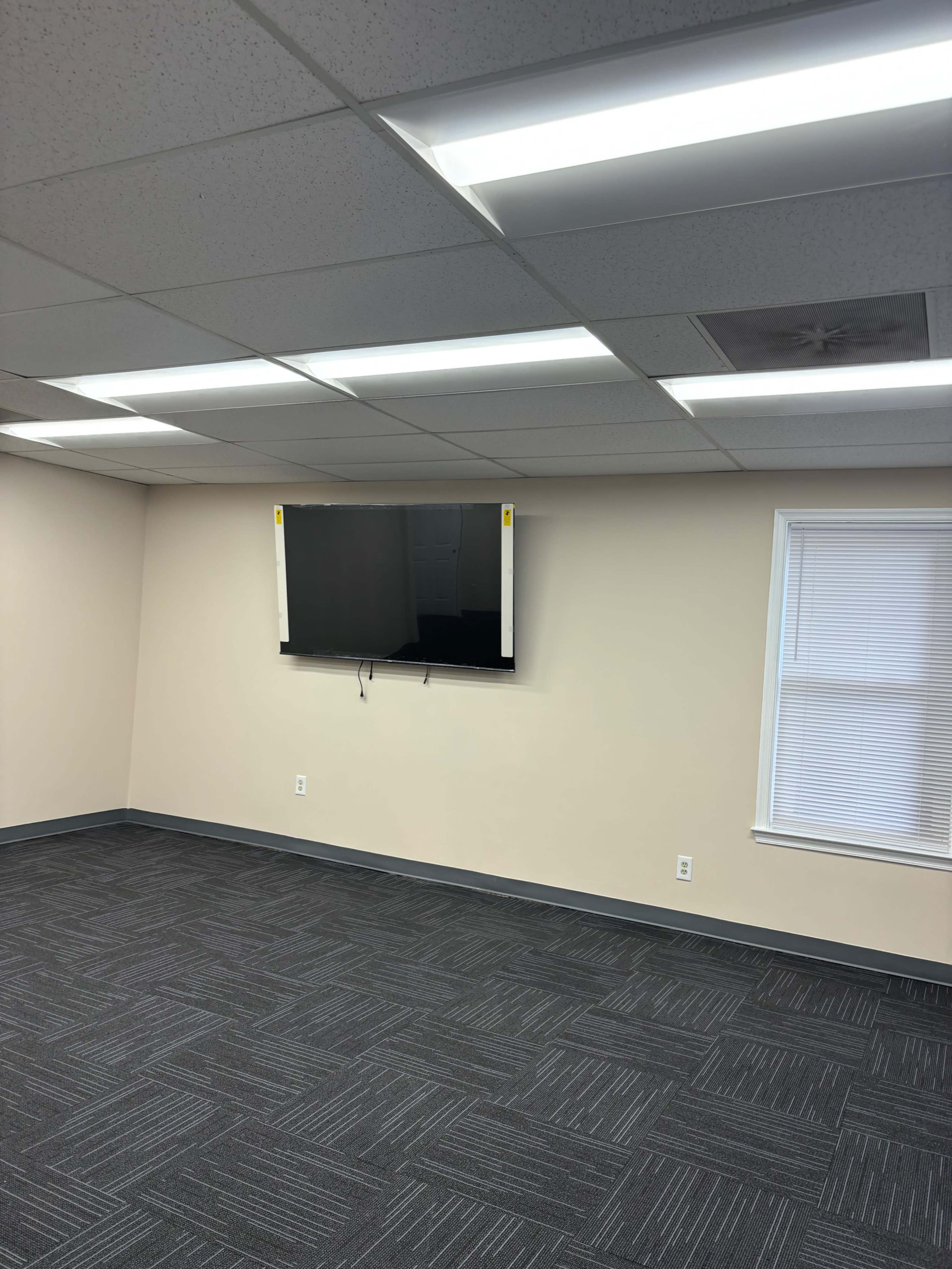 A flat-screen TV is mounted on a light-colored wall in a room with a gray carpet and fluorescent ceiling lights.