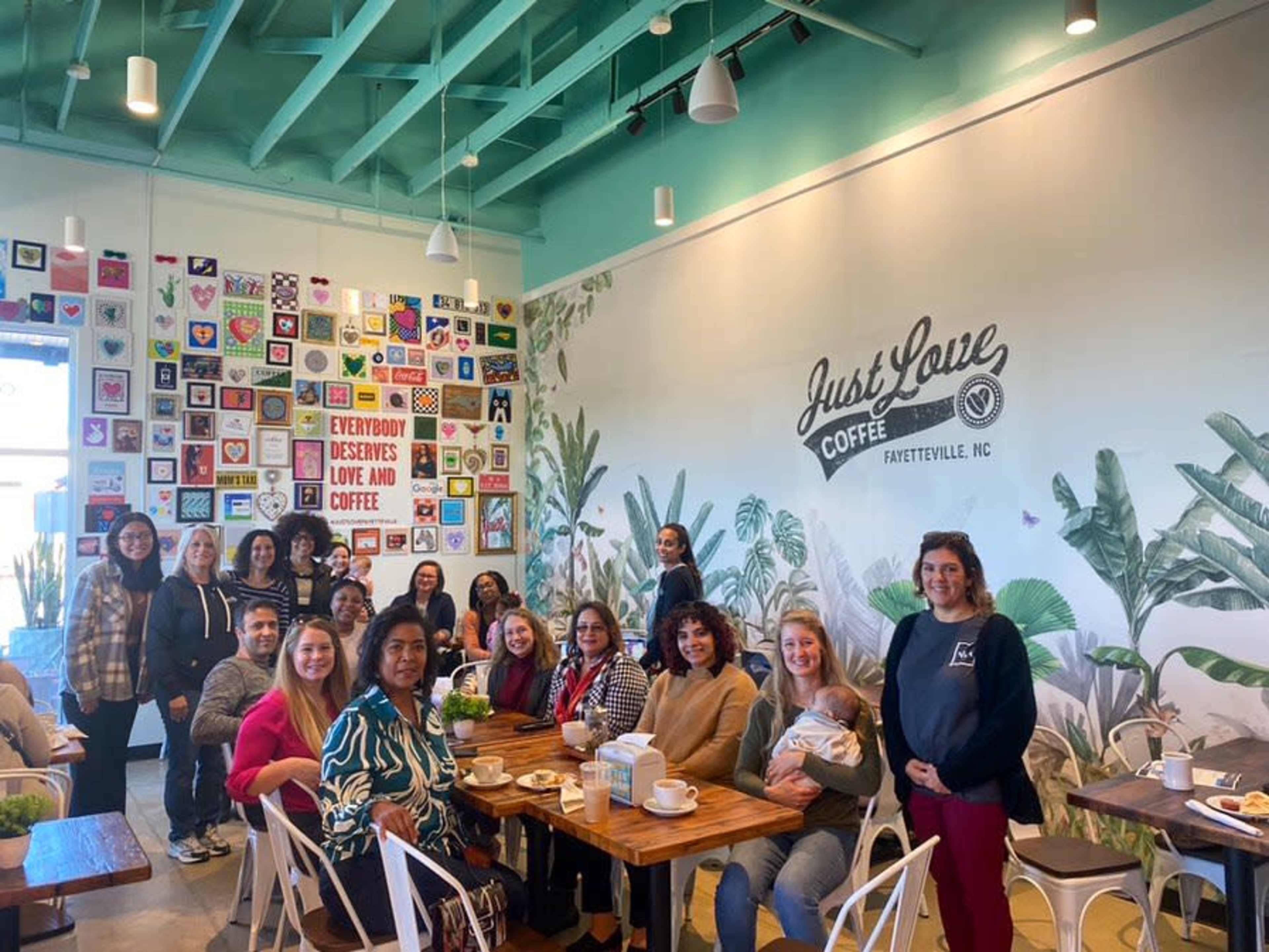 A group of twenty women gathers around tables in a brightly decorated coffee shop, with a colorful wall art display in the background.
