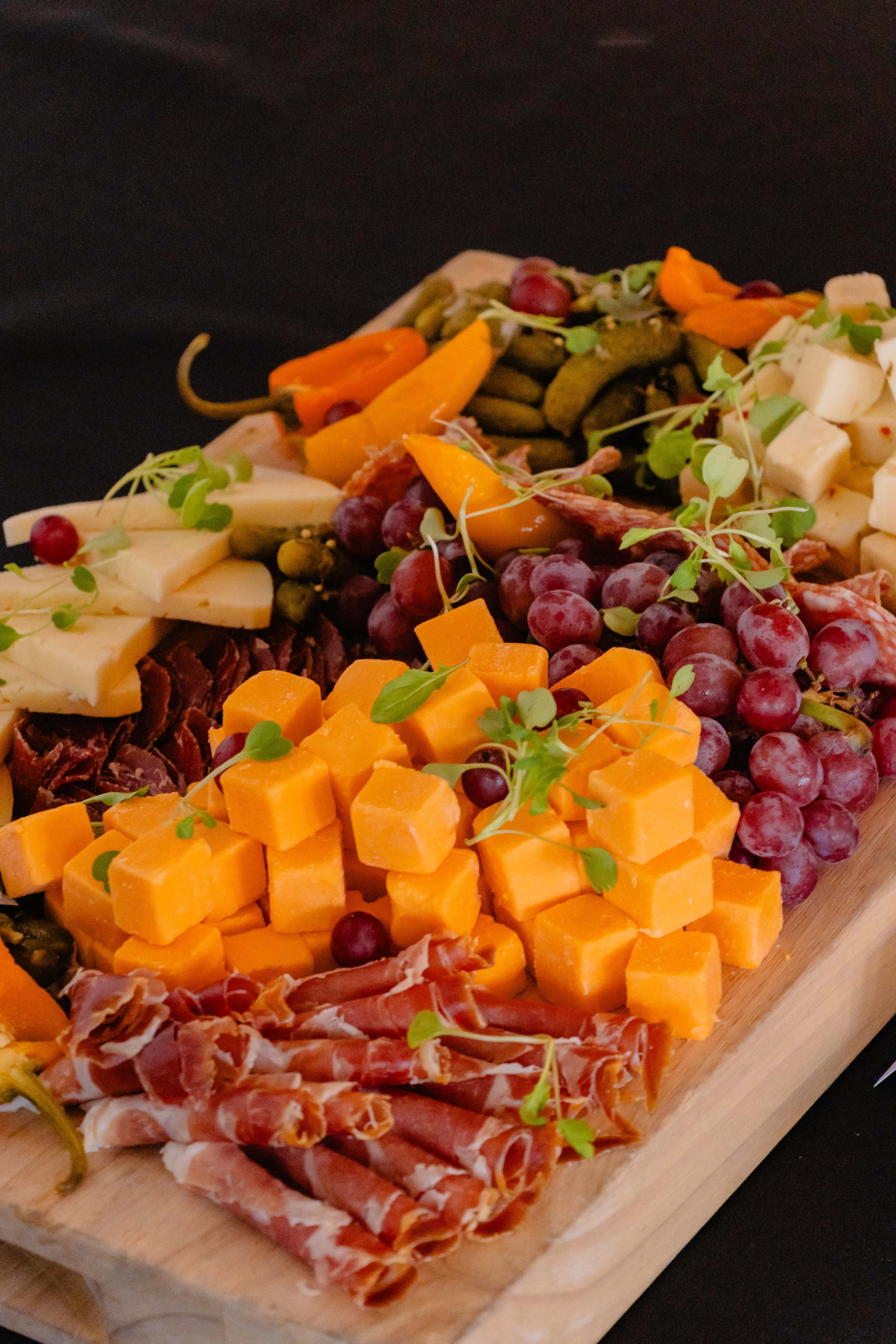 A wooden platter displays a variety of cheeses, cured meats, grapes, and pickled vegetables.