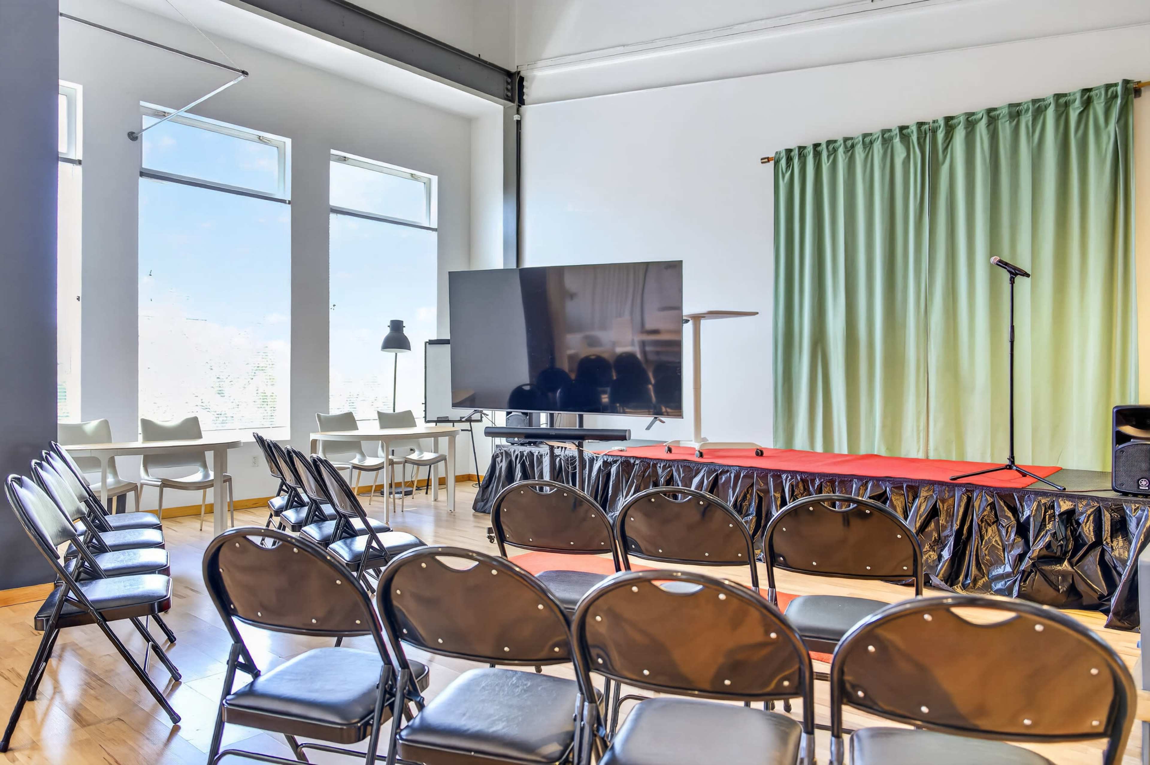 The image shows a spacious room set up for an event, featuring rows of black chairs facing a stage with a microphone and a large screen.