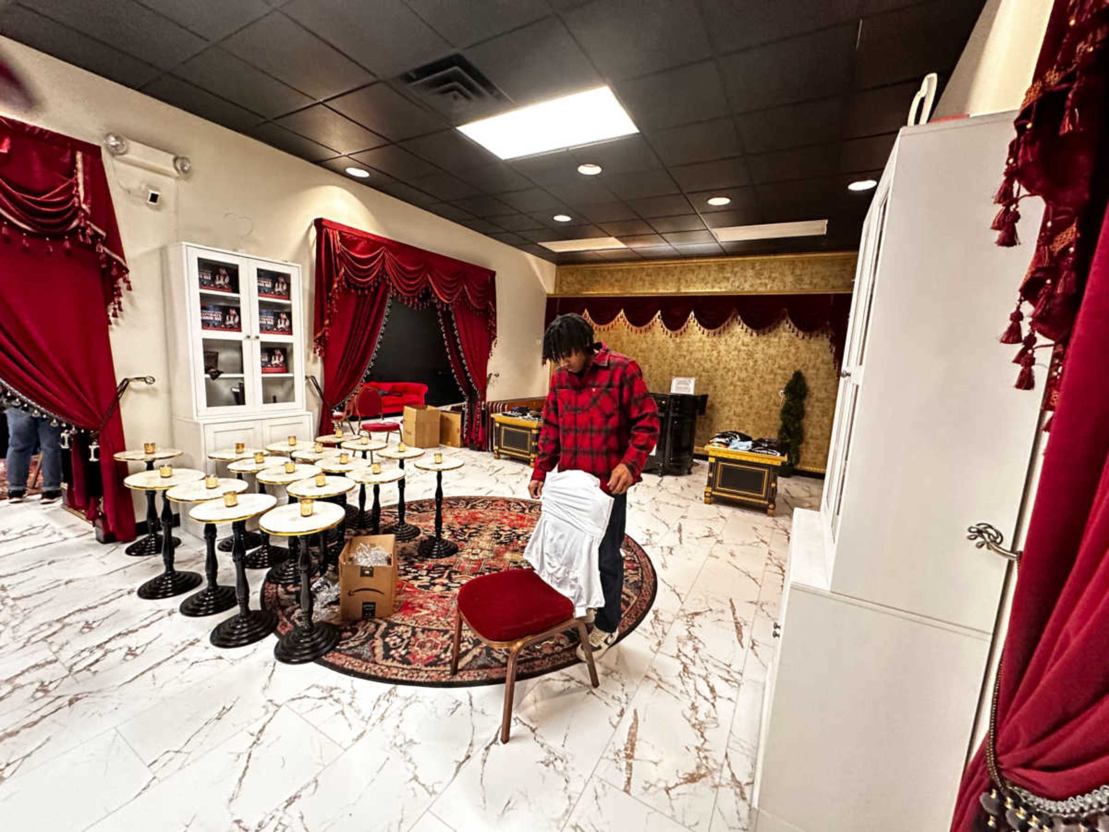A person wearing a red checkered shirt is standing in a room with decorated curtains, a circular rug, and several small tables arranged around.