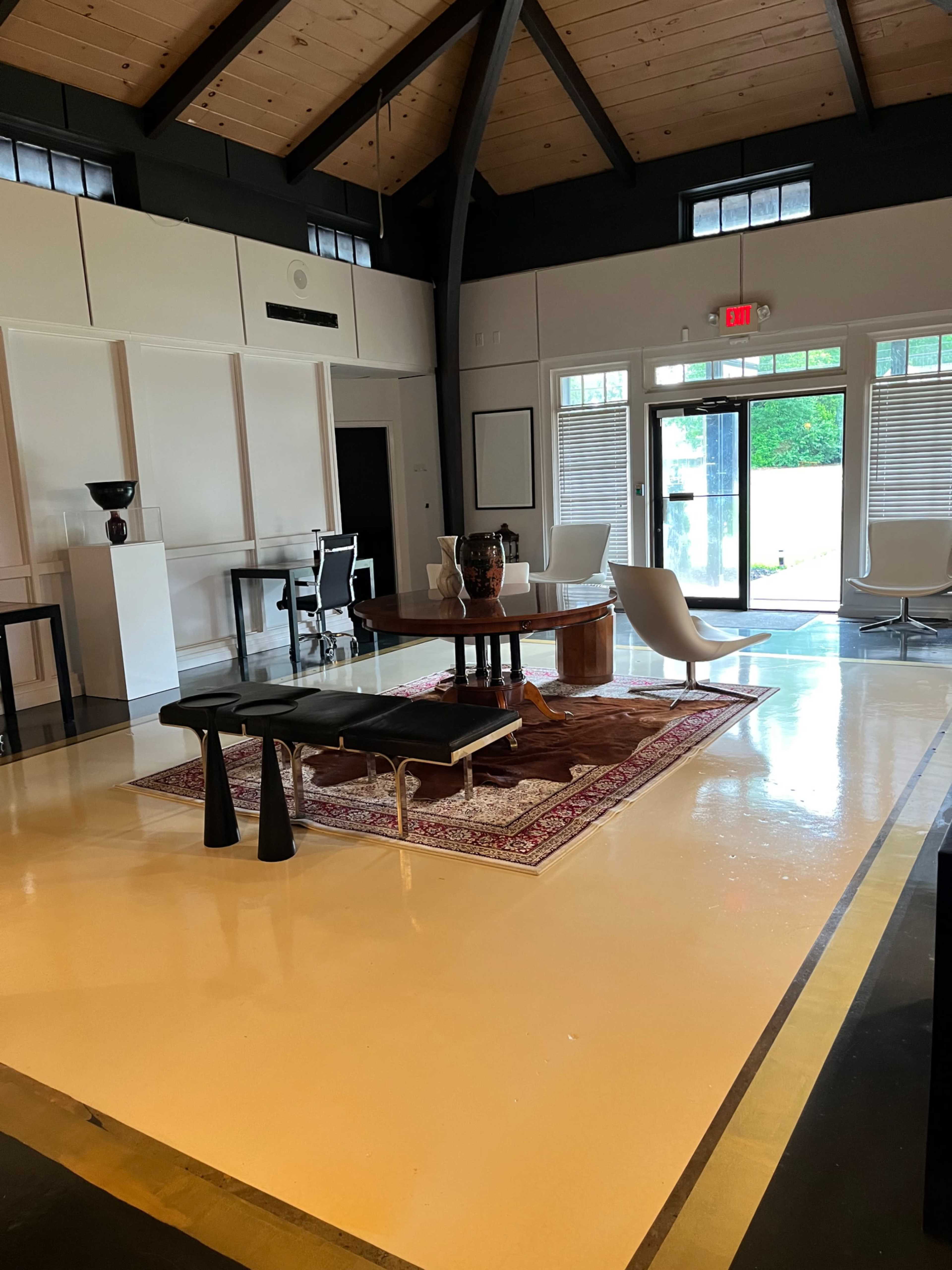 The image shows a modern interior space with a large round table at the center, surrounded by various seating options, and large windows letting in natural light.
