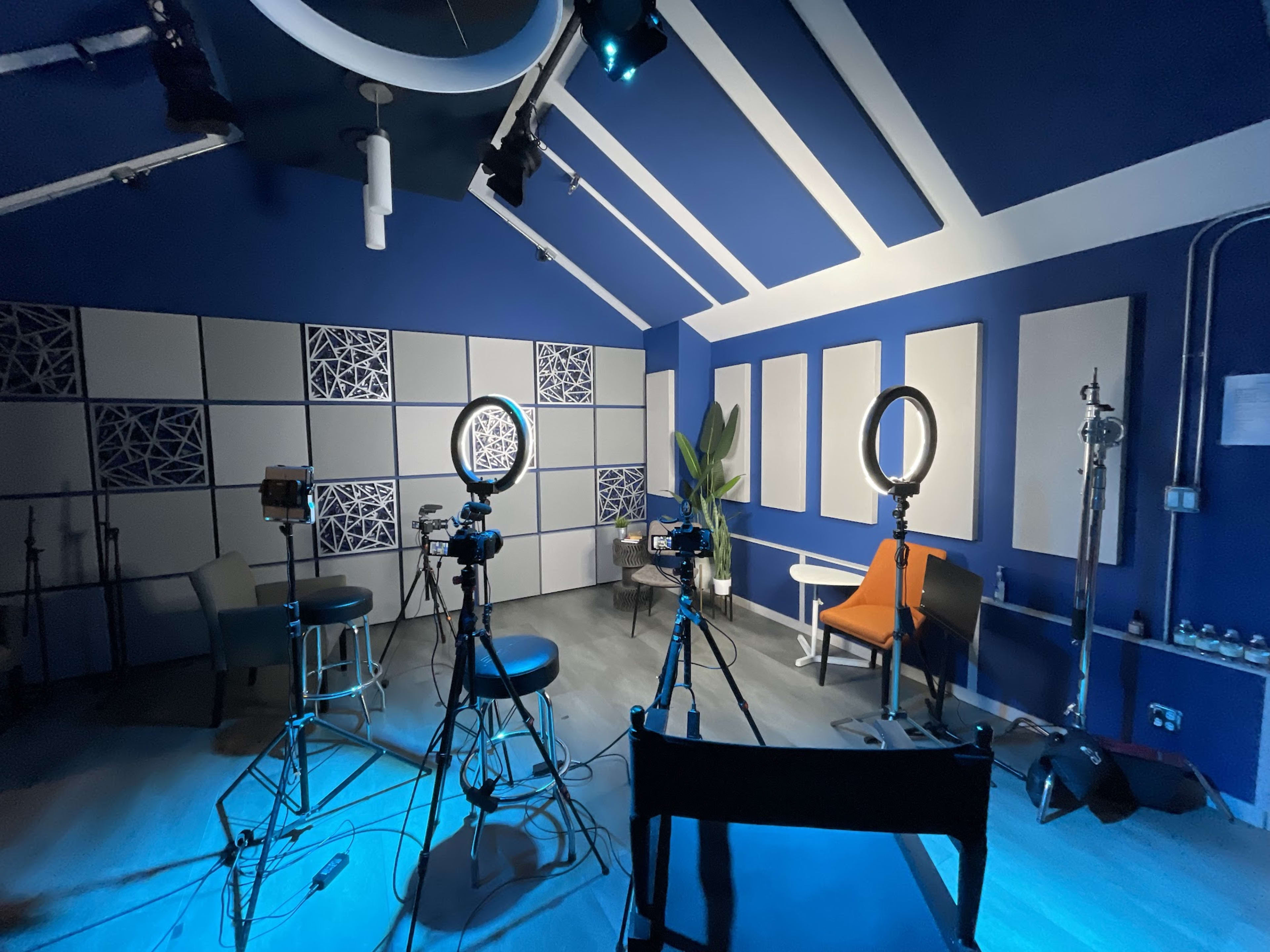A filming studio with several cameras, lighting equipment, and a few chairs arranged against a blue wall.