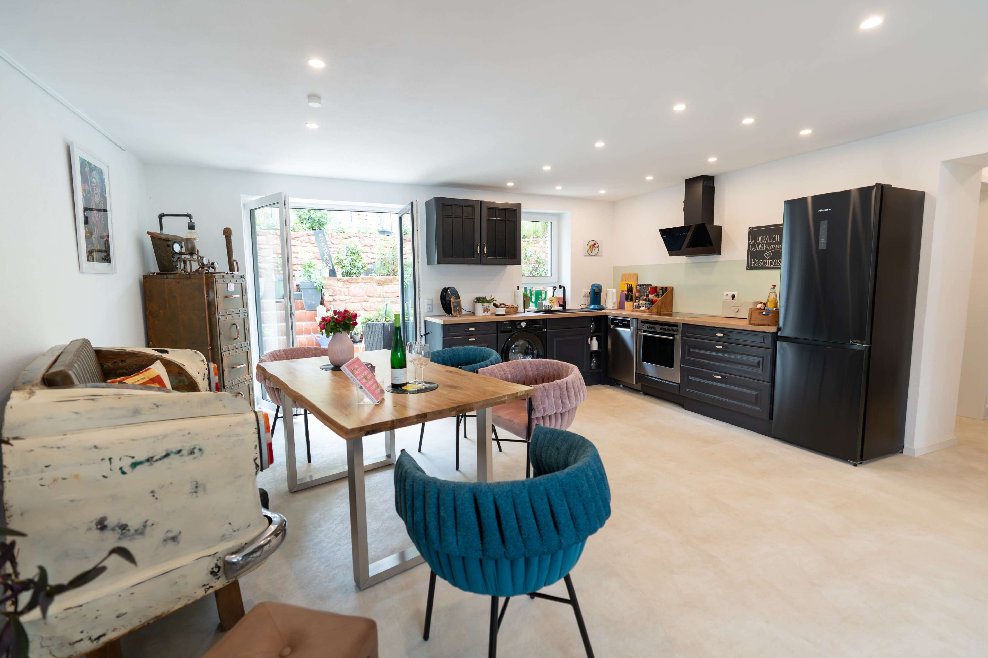 The image features a modern kitchen with a dining table surrounded by colorful chairs, black cabinetry, and an open doorway leading to an outdoor area.