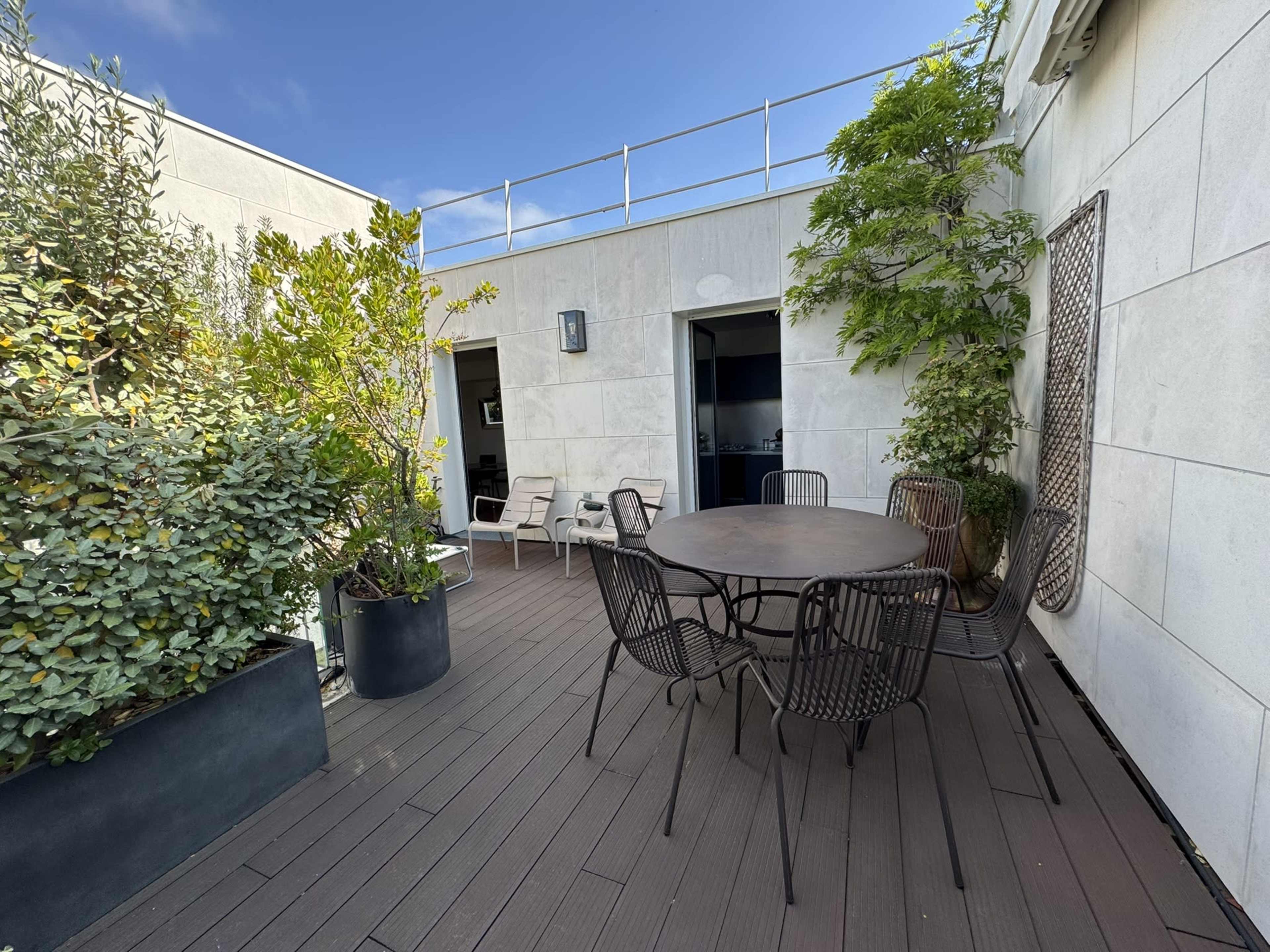 The image shows a rooftop terrace featuring a round table surrounded by chairs, planters with greenery, and two doorways leading into the interior.