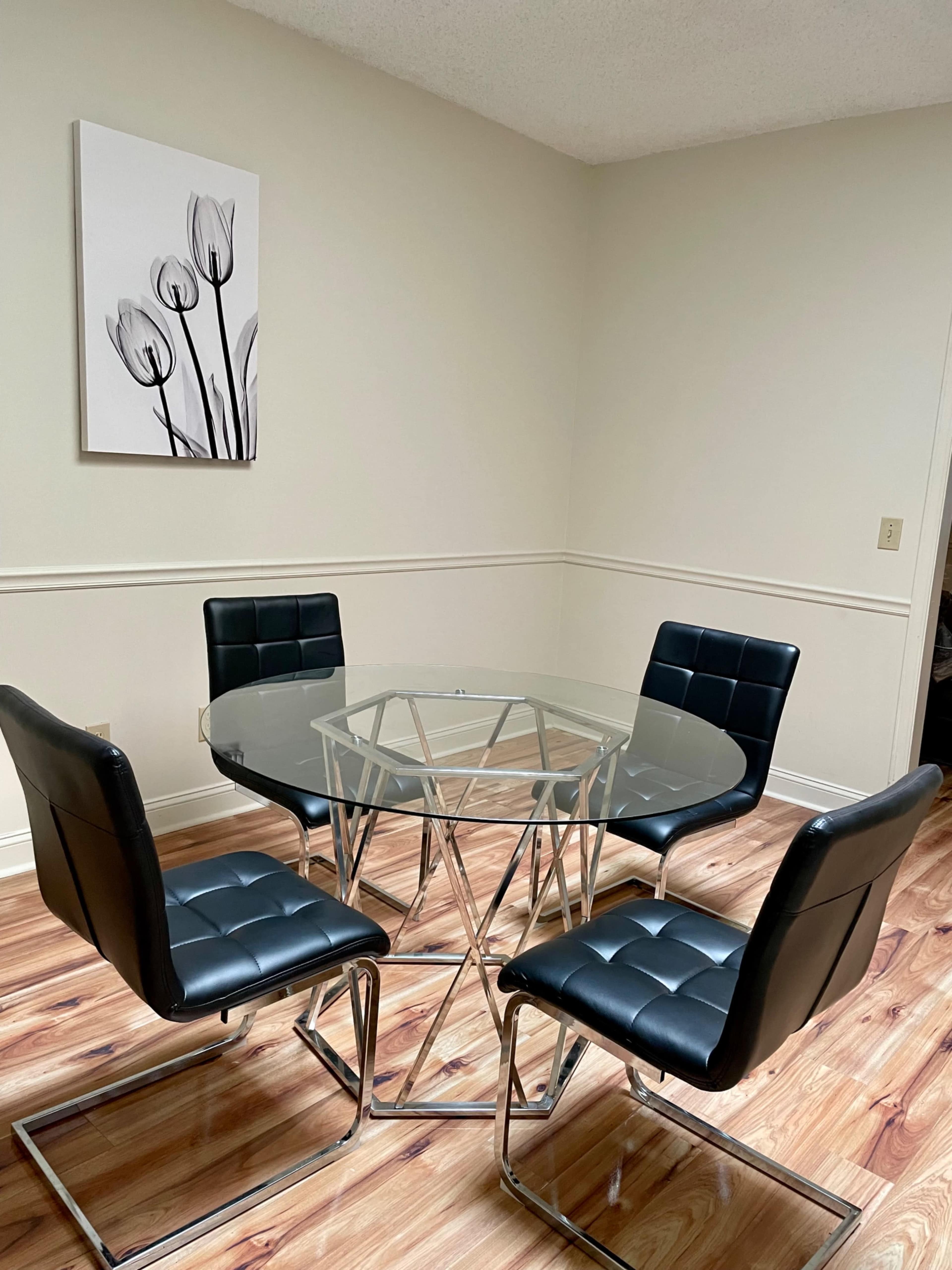 A modern glass dining table surrounded by four black upholstered chairs in a minimalist room with wooden flooring.