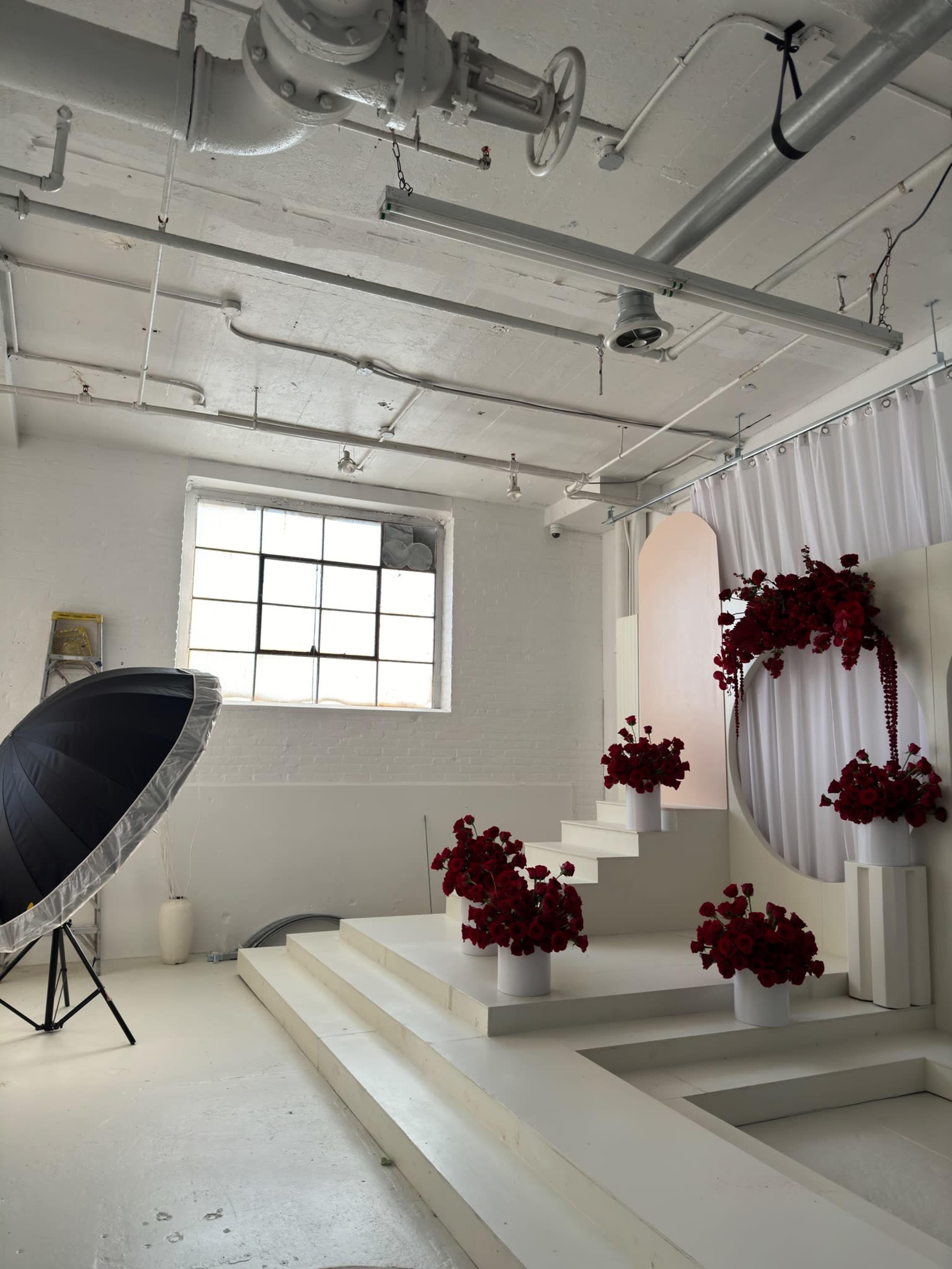 A bright studio interior featuring a set of white steps adorned with red flowers, alongside large photography equipment and a window letting in natural light.