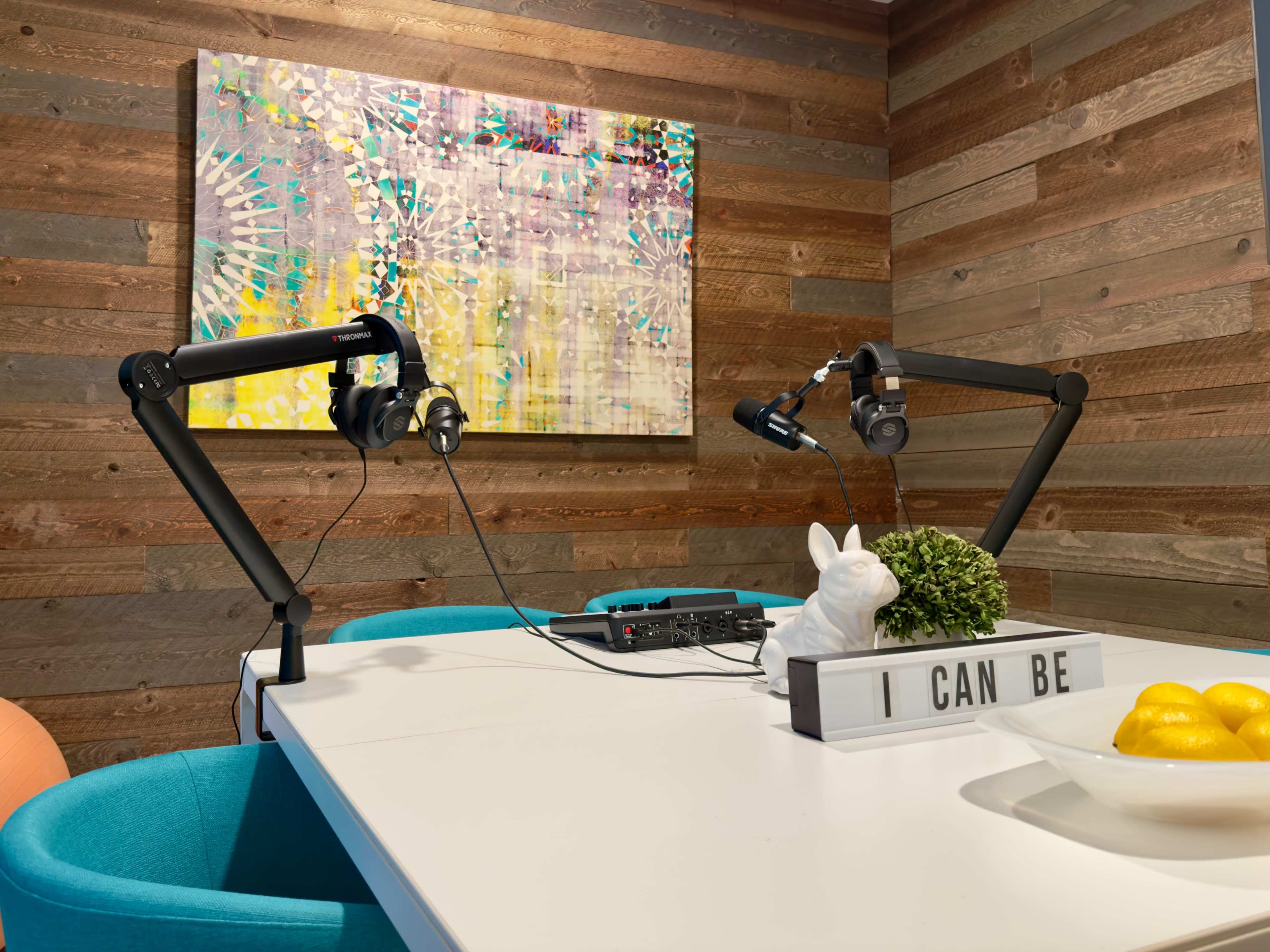 A brightly colored abstract painting hangs on a wooden wall above a white table with microphones and audio equipment set up for recording, alongside a decorative rabbit figurine and a bowl of lemons.