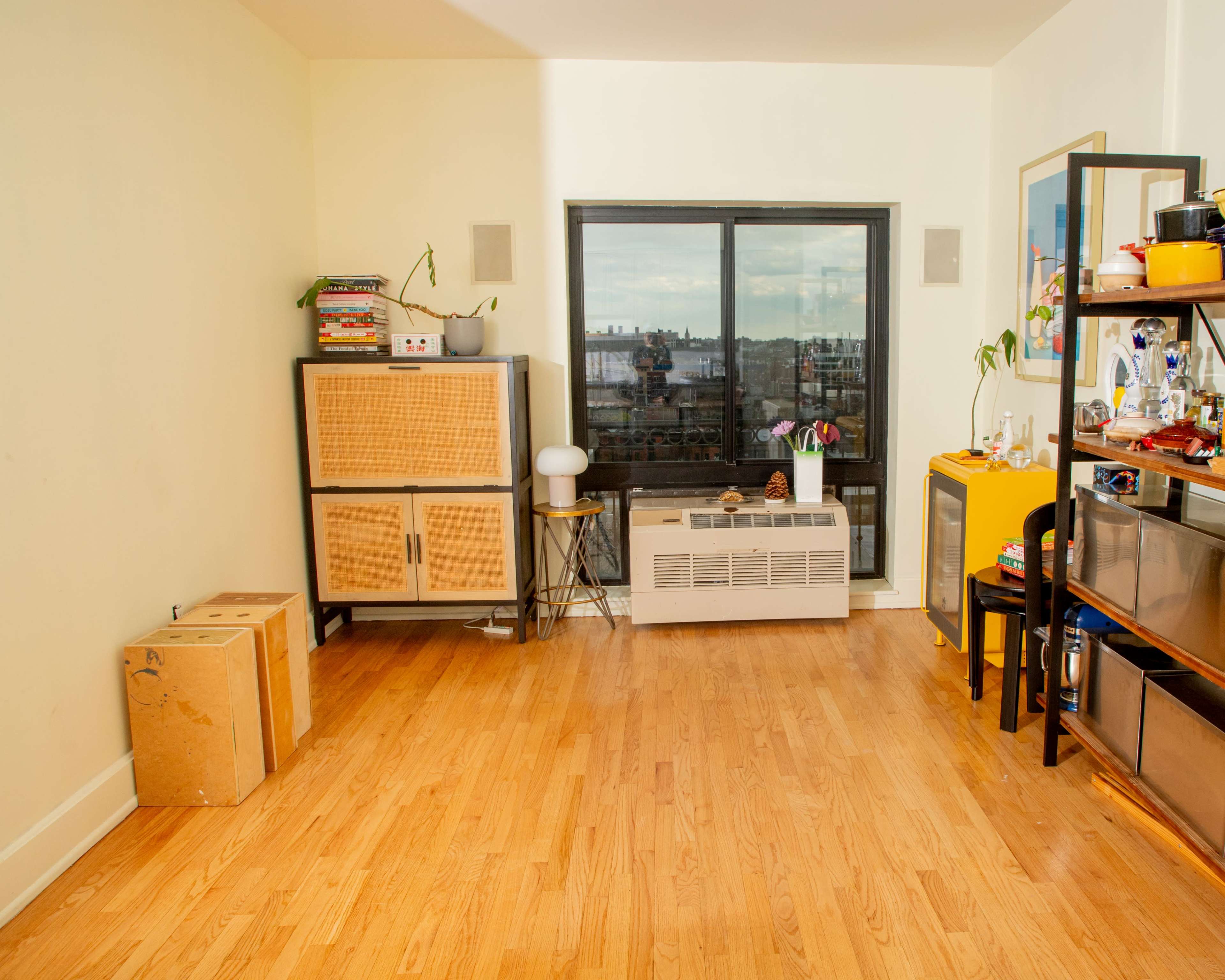 The image shows a small, sparsely furnished room with wooden flooring, featuring a cabinet, a television stand with various items, and a large window overlooking a cityscape.