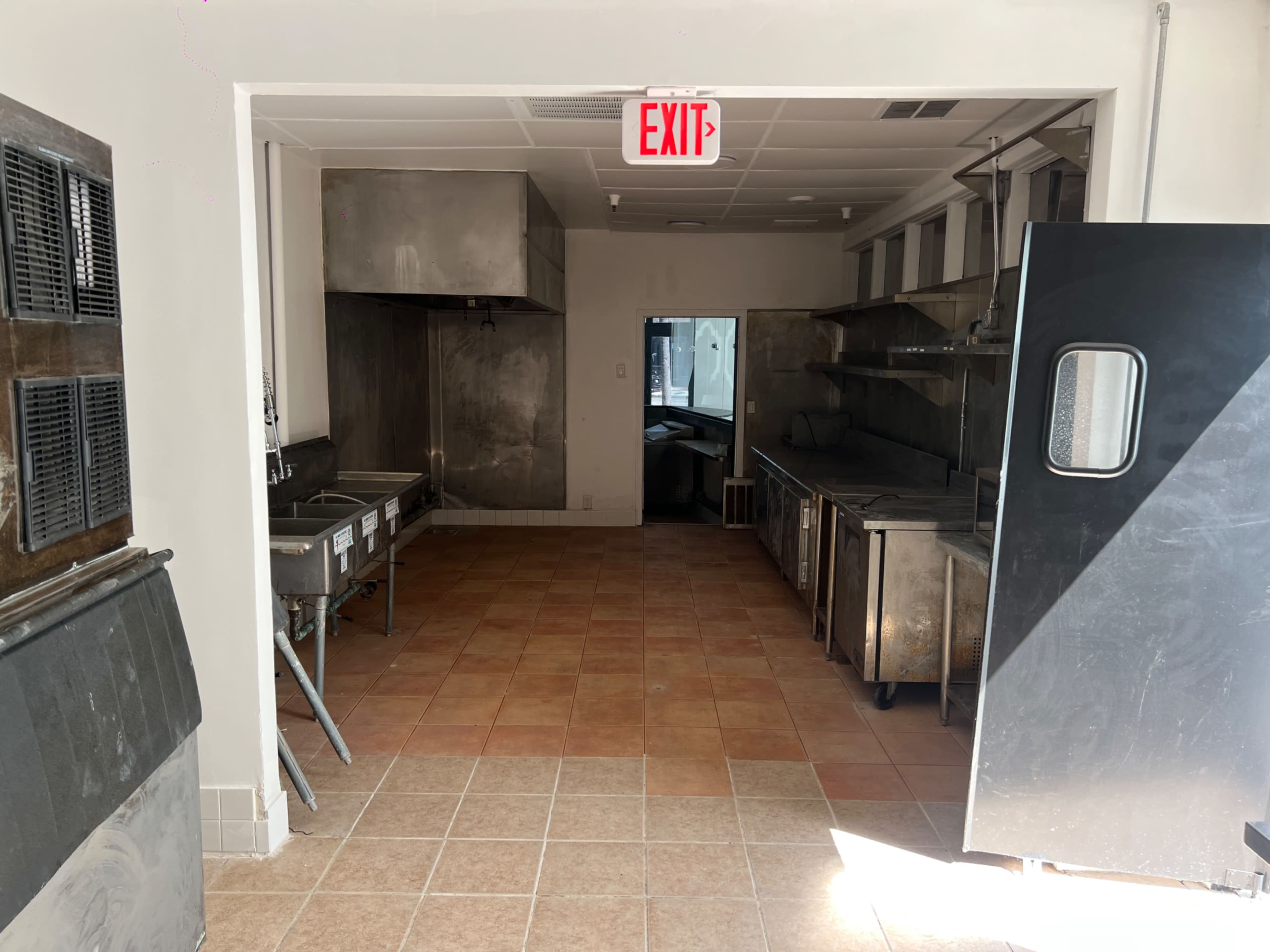 The image shows an empty commercial kitchen with stainless steel appliances, a tiled floor, and an exit sign visible above a doorway.