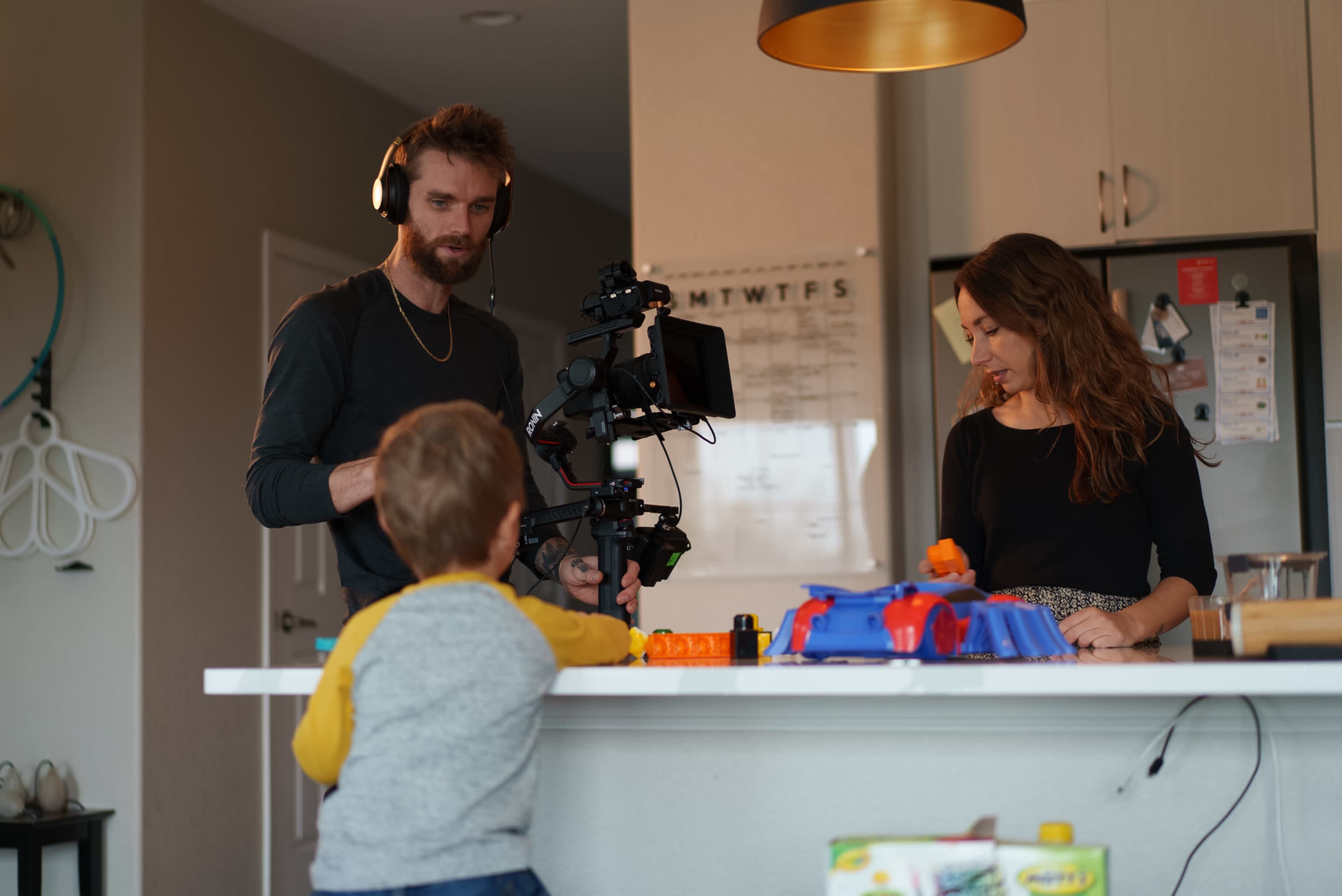 A man with headphones operates a camera on a stabilizer while a woman watches in a modern kitchen, as a child observes from the countertop.