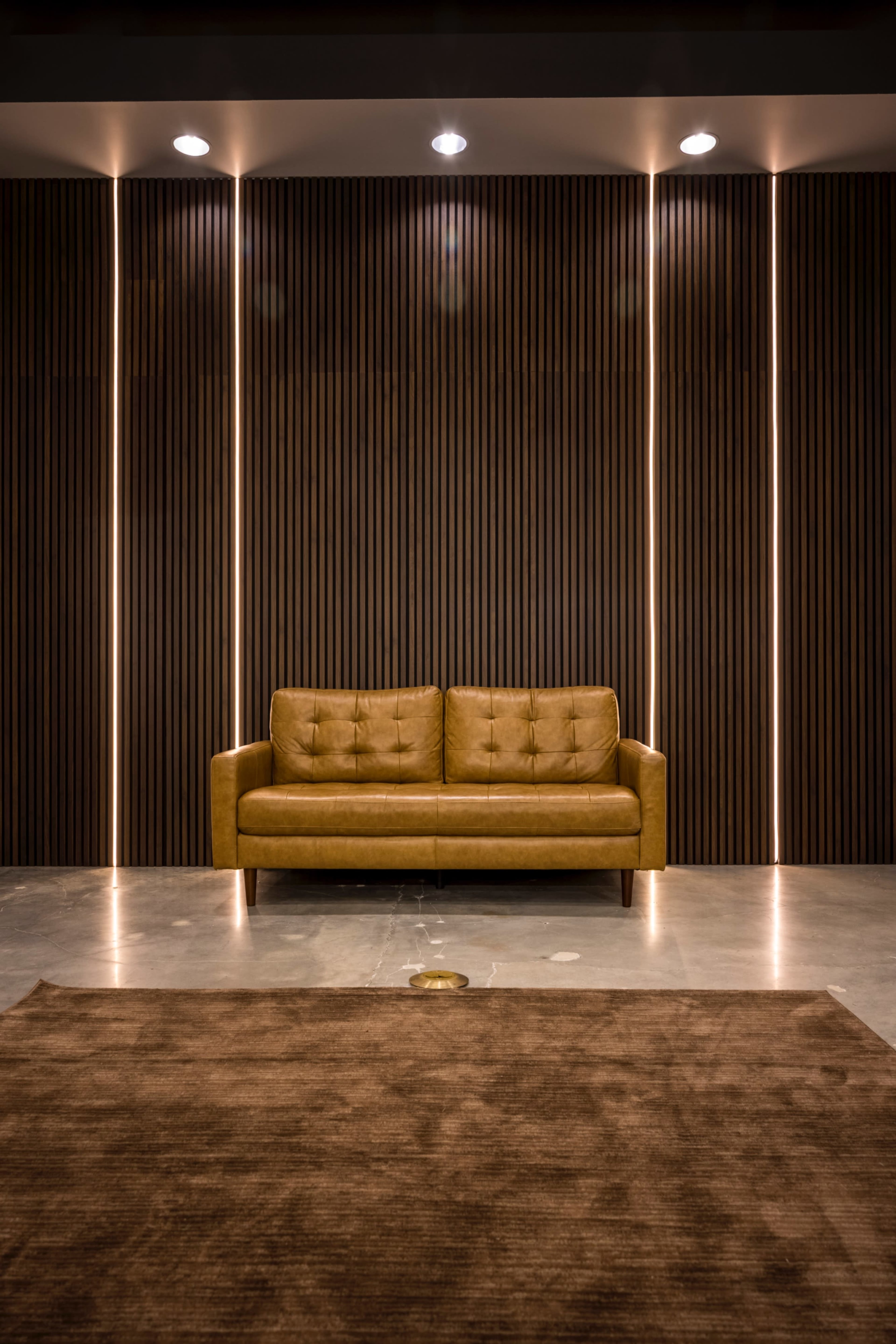 A sleek, brown leather sofa is positioned against a wall with vertical wooden slats and soft lighting.