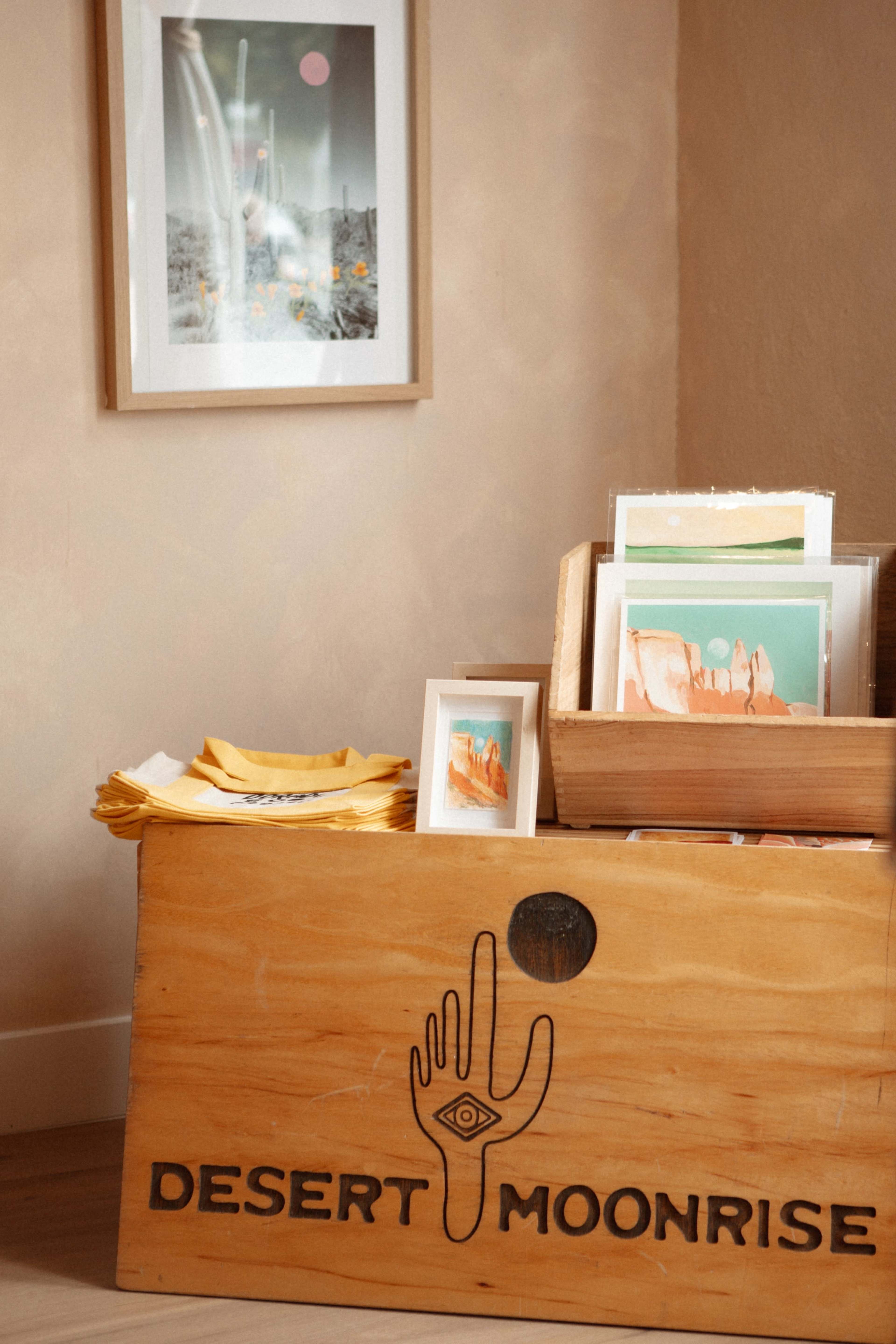 The image shows a wooden display box labeled "DESERT MOONRISE" filled with small artworks and photographs, next to a framed picture on the wall.