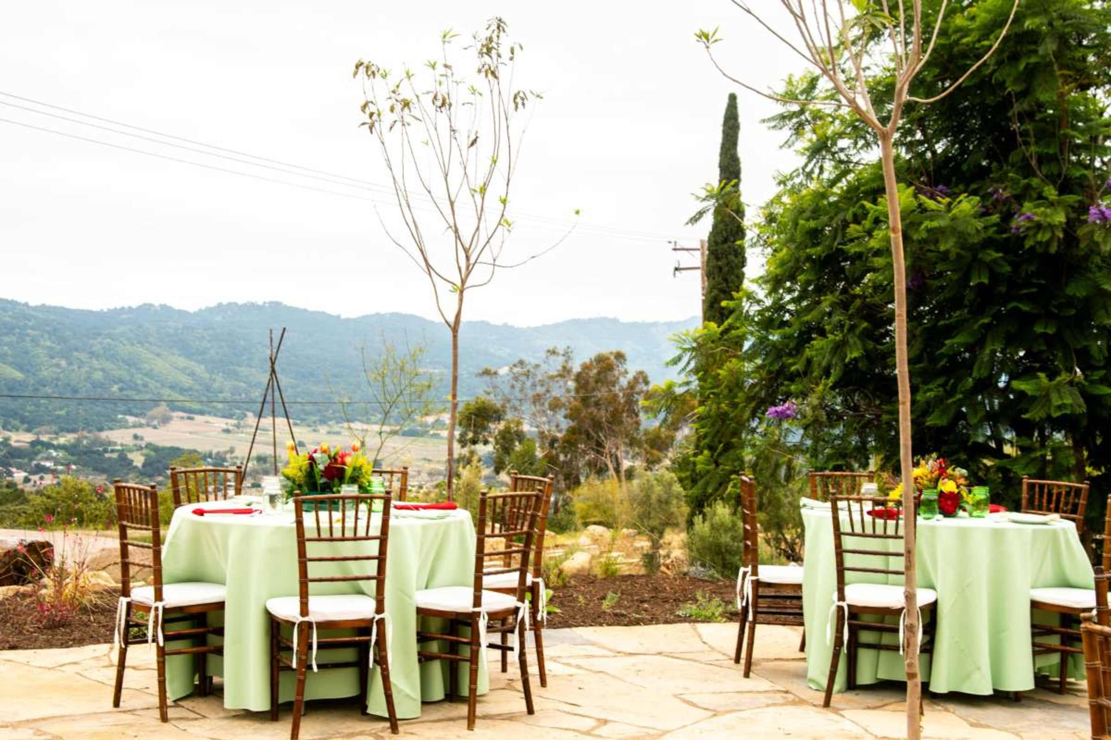 A landscaped outdoor venue features round tables with green tablecloths set for a gathering, overlooking a hilly landscape.