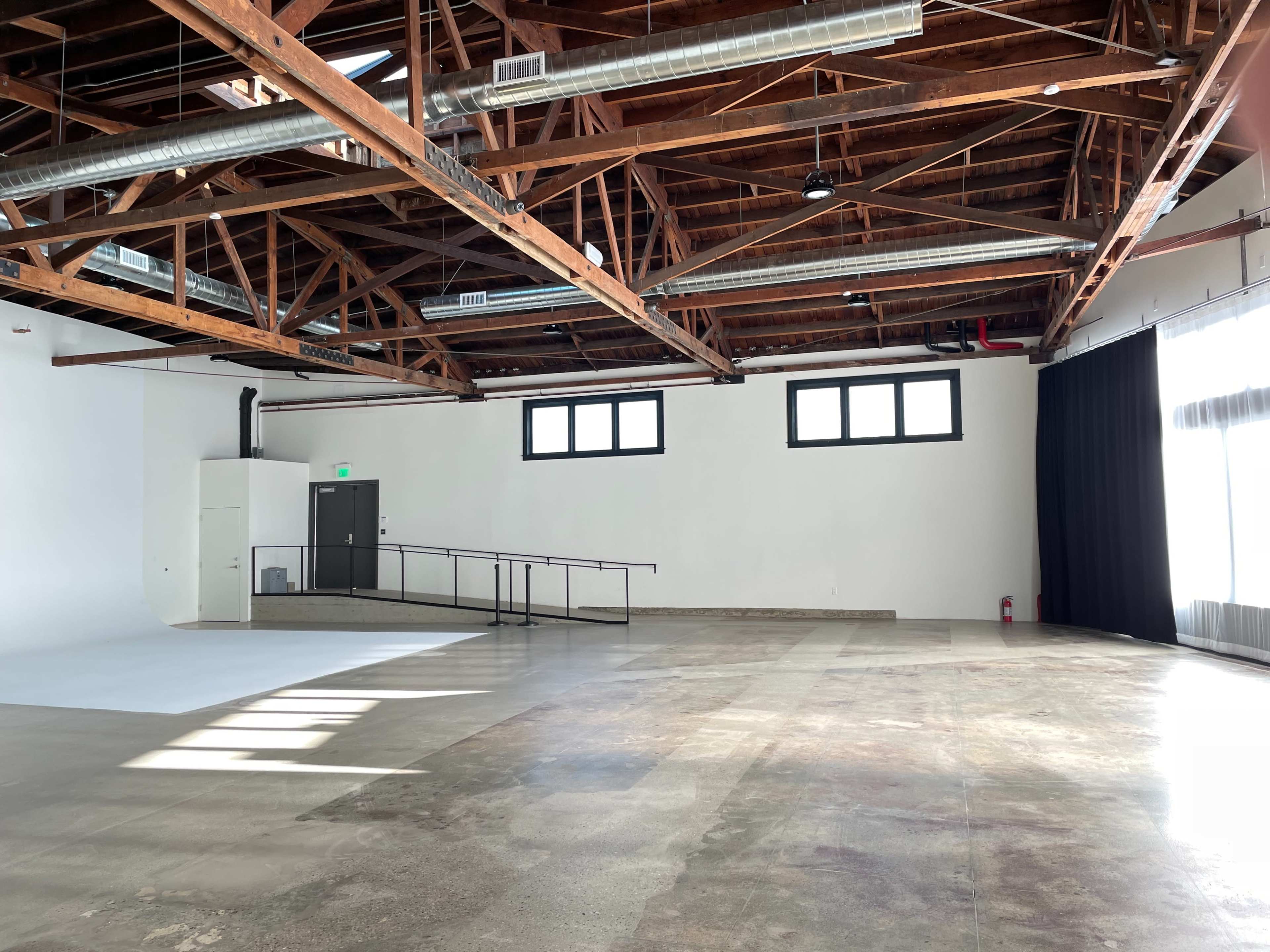 A spacious room with a wooden beam ceiling, concrete flooring, and large windows, featuring a white backdrop and a black curtain on one side.