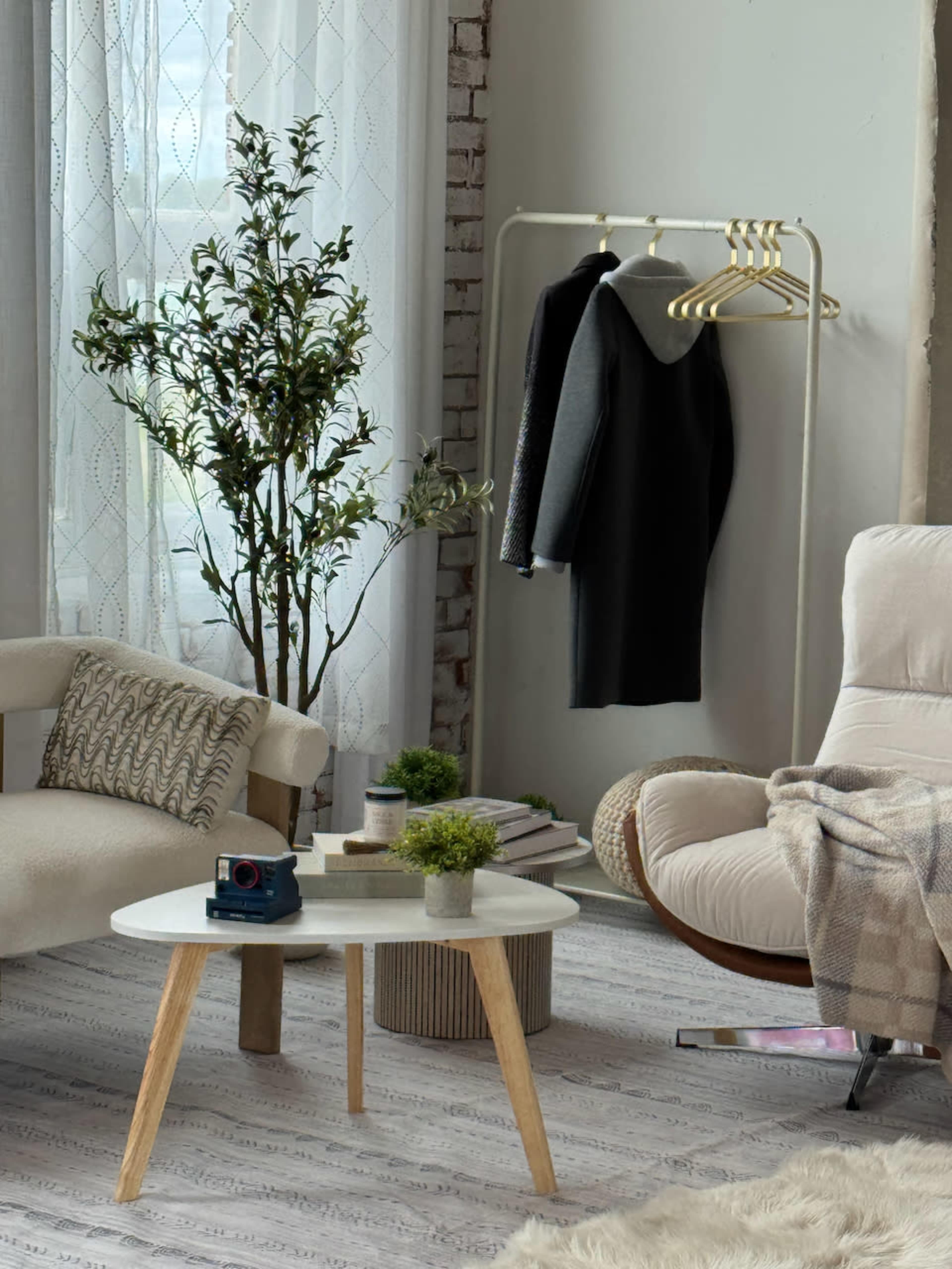 The image shows a cozy living space featuring a white round coffee table, a plant next to a clothing rack with garments, and a cream-colored armchair with a throw blanket draped over it.