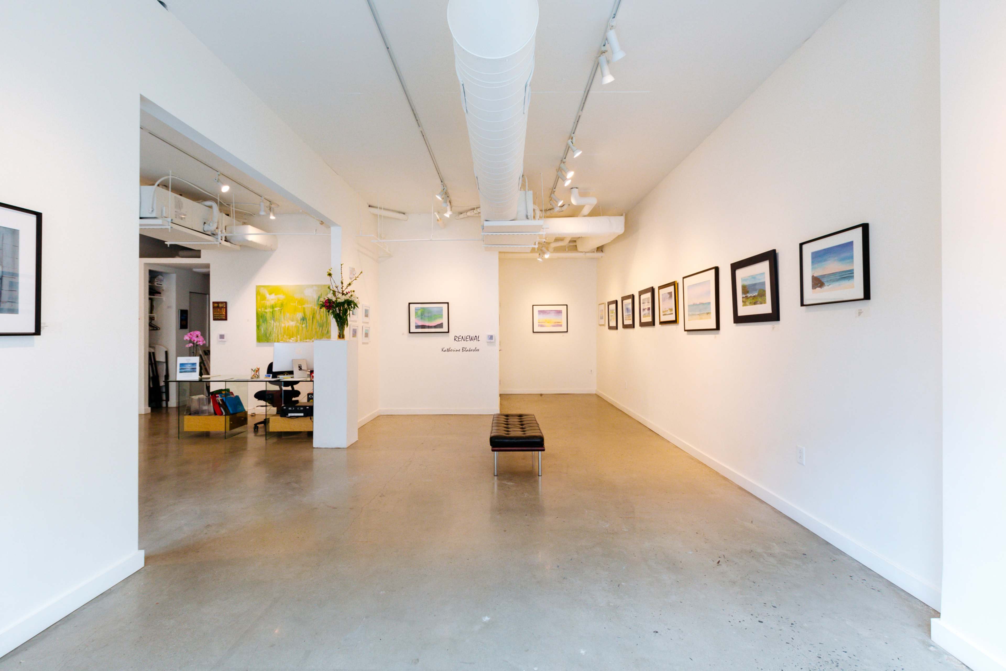 An art gallery with framed photographs displayed on white walls, a seating bench in the center, and a workspace area visible in the background.