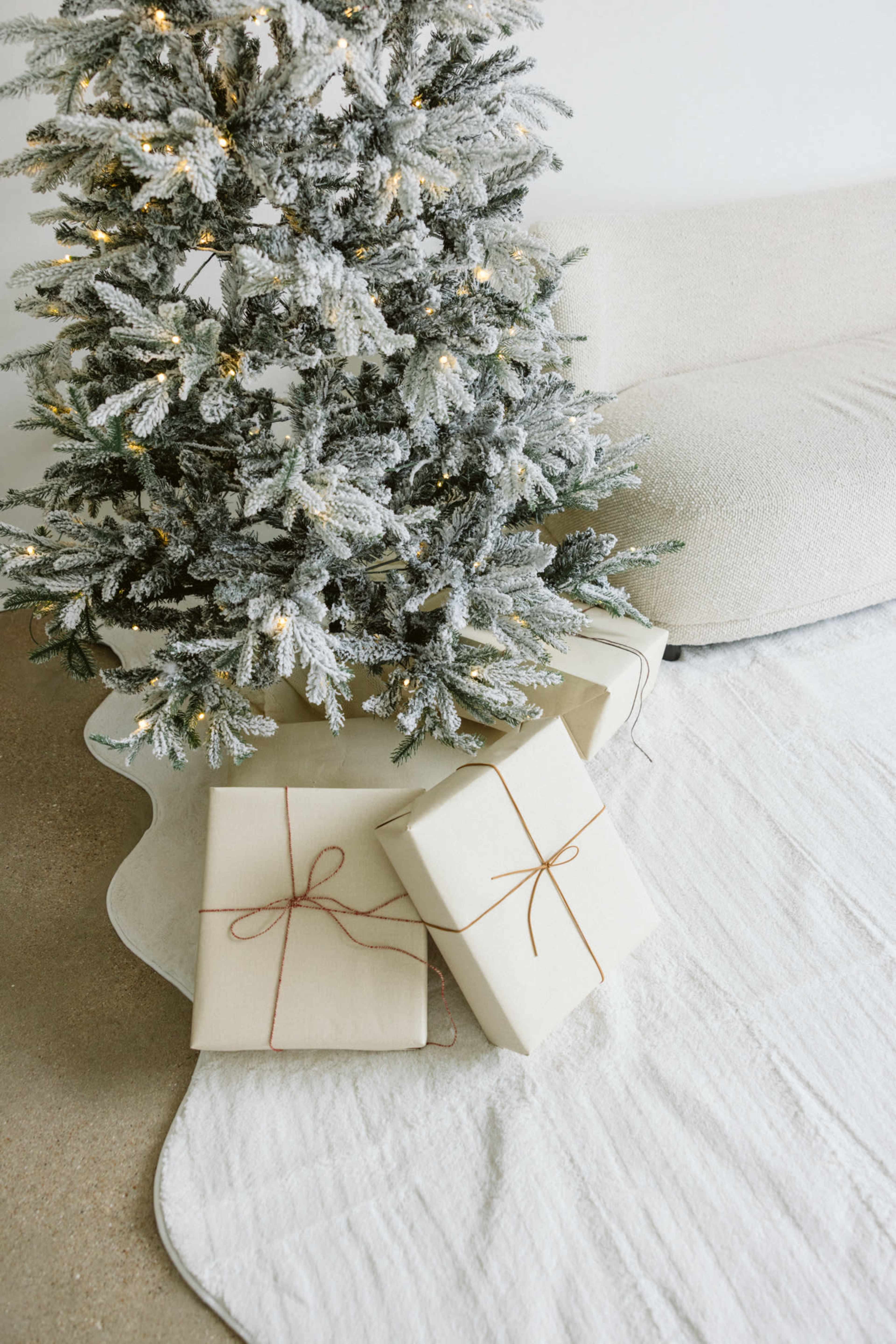 A frosted Christmas tree stands beside a couch, with two neatly wrapped presents on the white rug beneath it.
