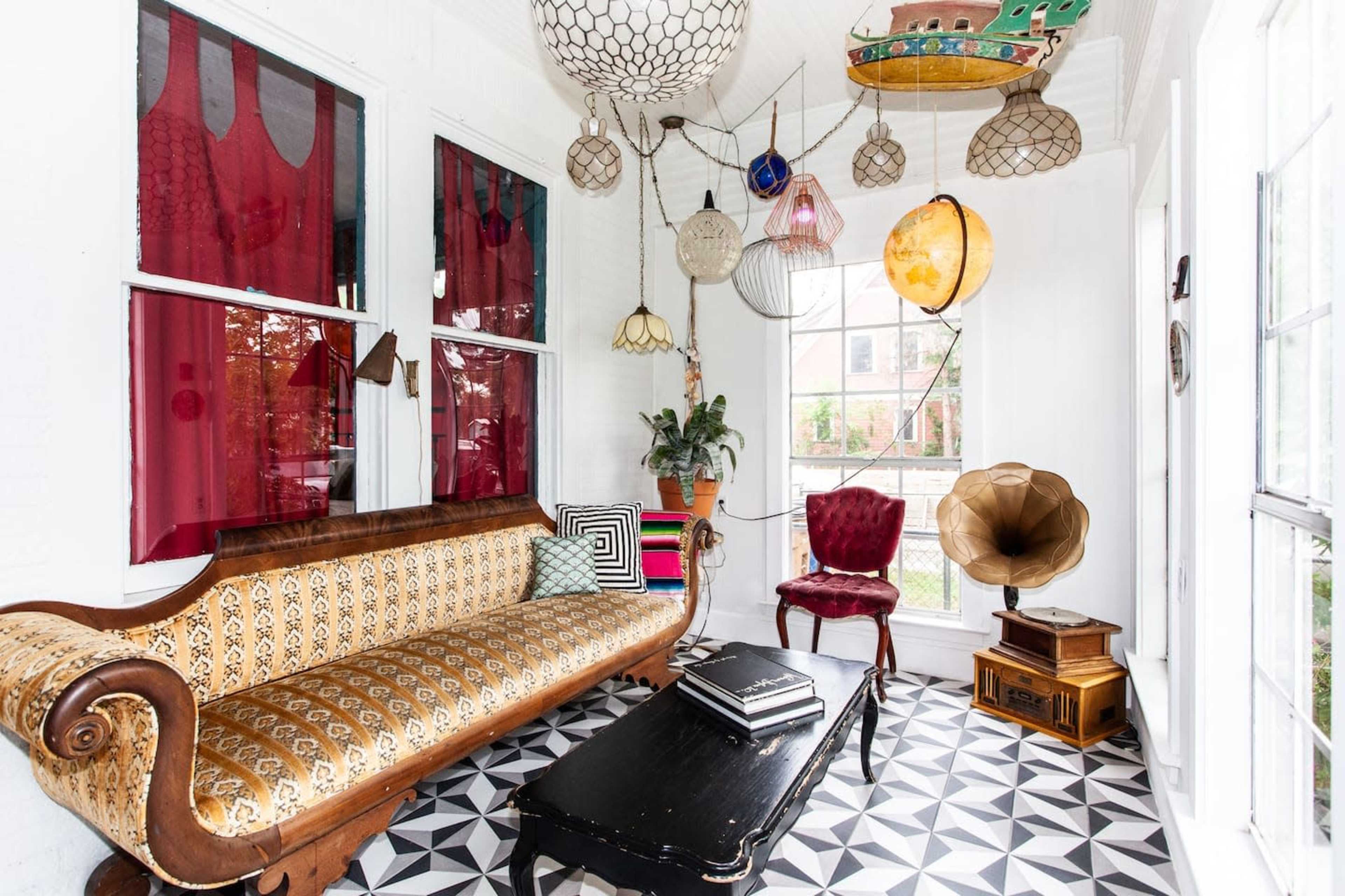 A brightly lit room featuring a vintage brown sofa, a round wooden table, a red velvet chair, and various decorative hanging light fixtures.
