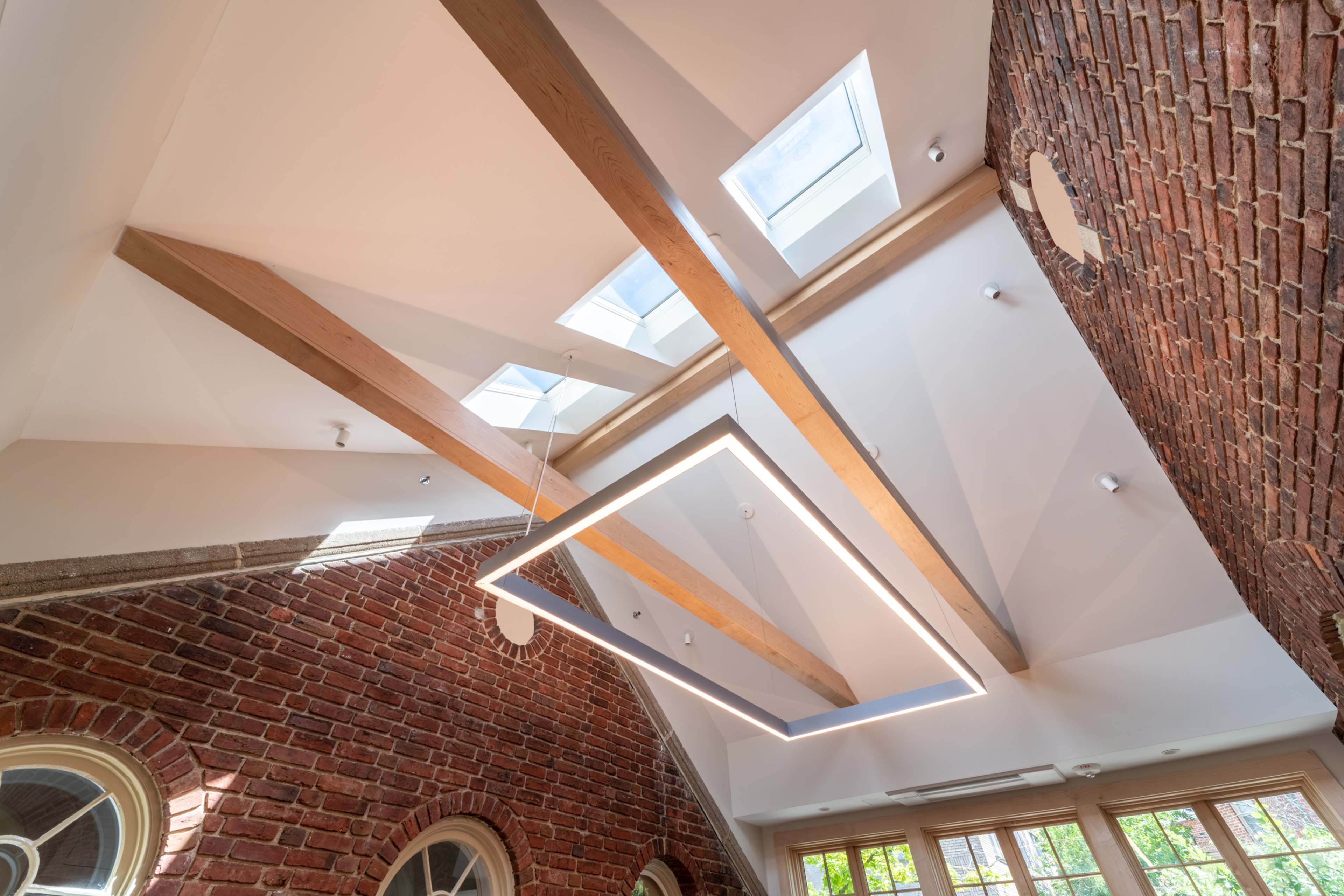 A modern ceiling with exposed wooden beams and skylights in a space featuring a red-brick wall.