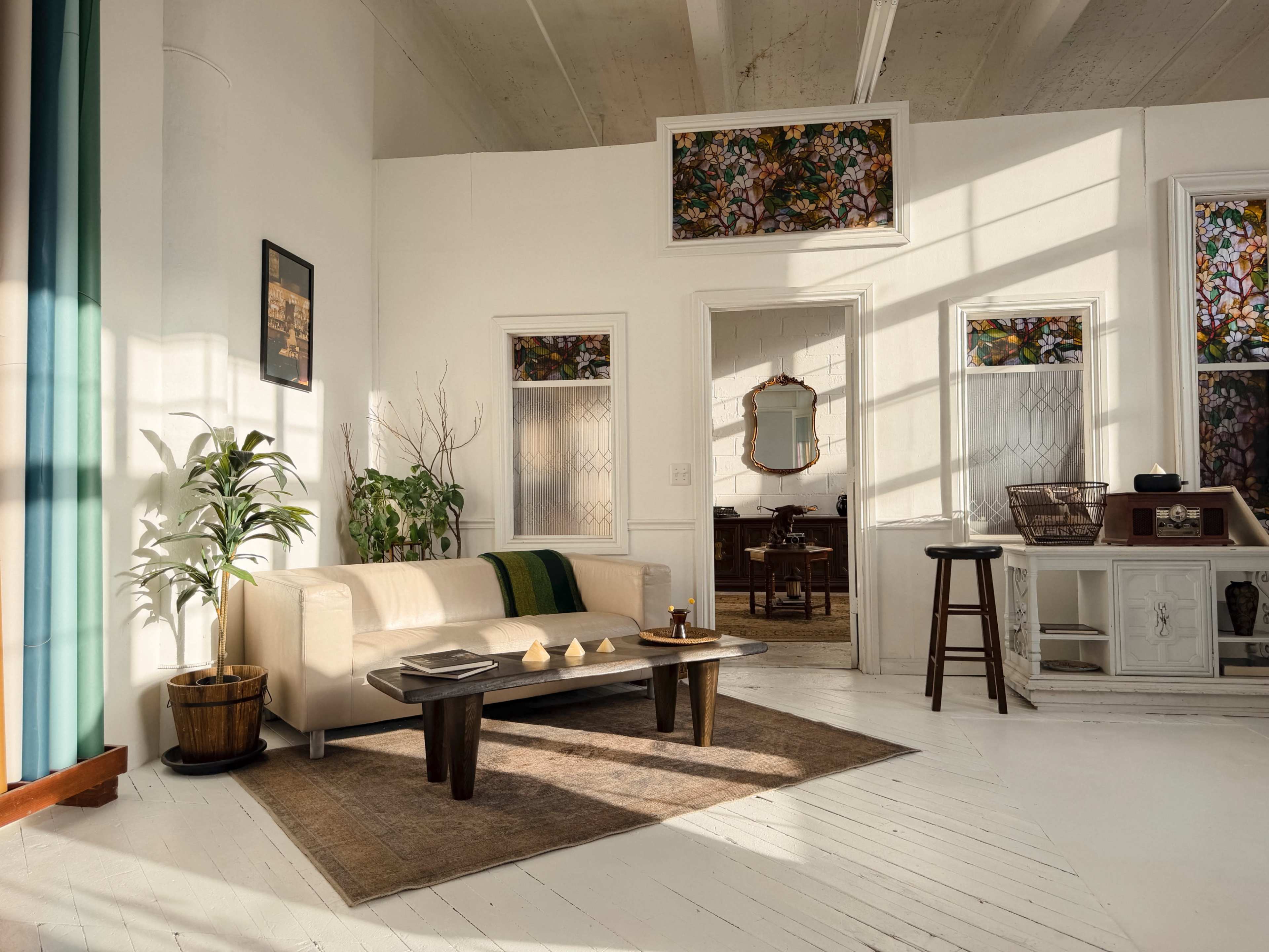 The image depicts a bright, modern living room with a sofa, coffee table, and decorative plants, featuring large windows that fill the space with natural light.