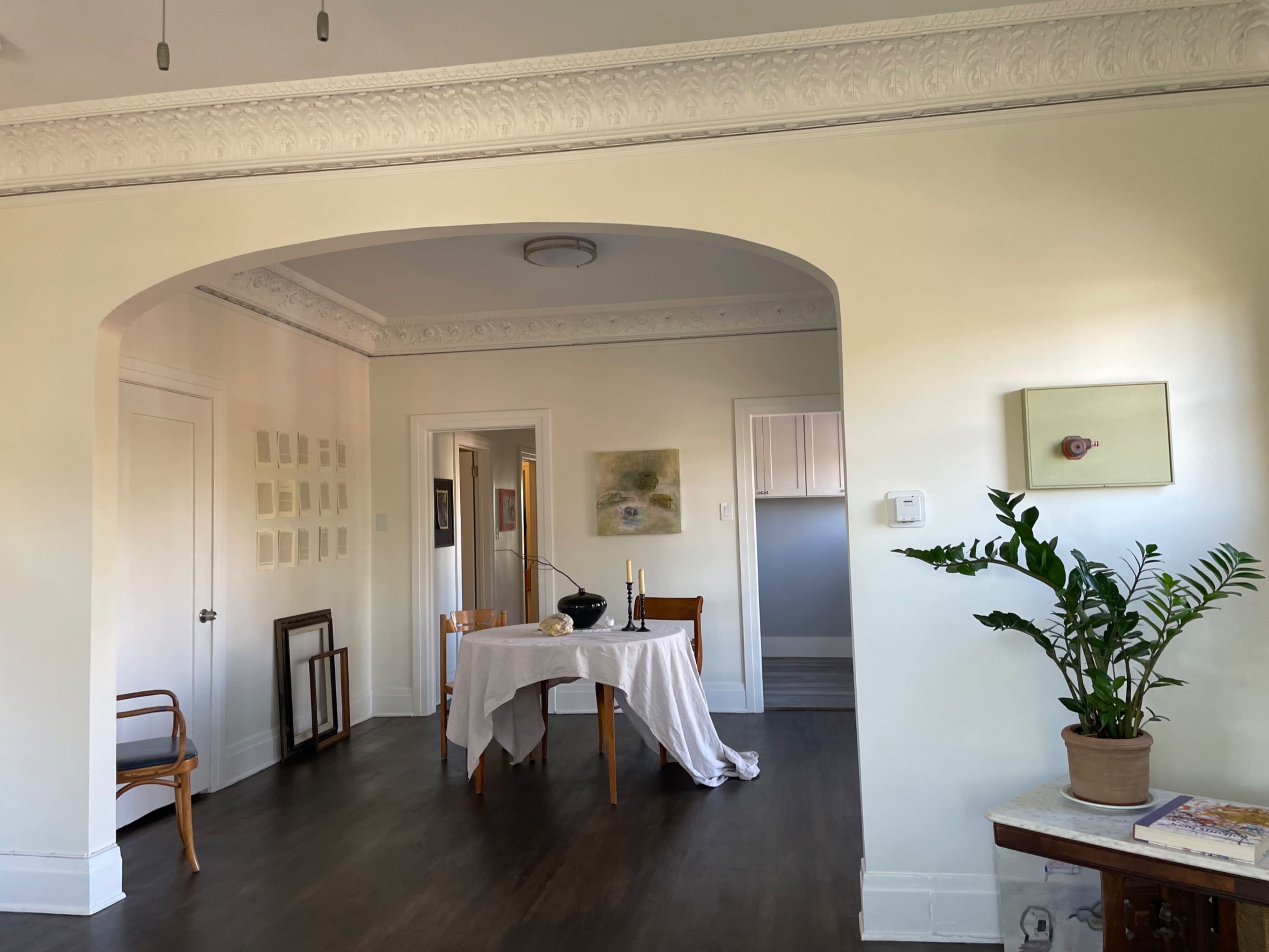 A dining table with a vase in the center is set in an open room, featuring a potted plant and framed artworks on the walls.