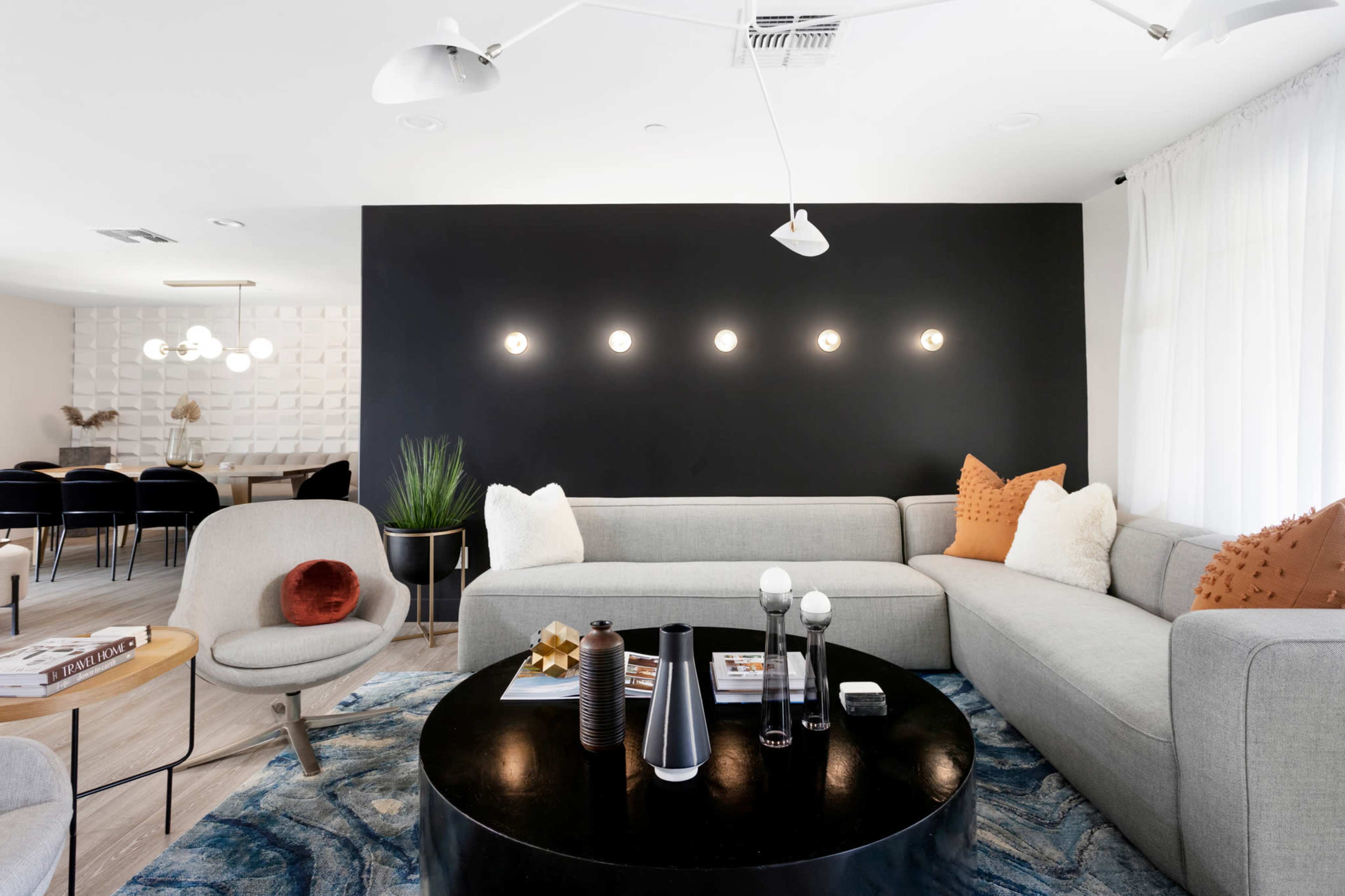 A modern living room features a gray sectional sofa, a round black coffee table, and a dark accent wall illuminated by five round lights.