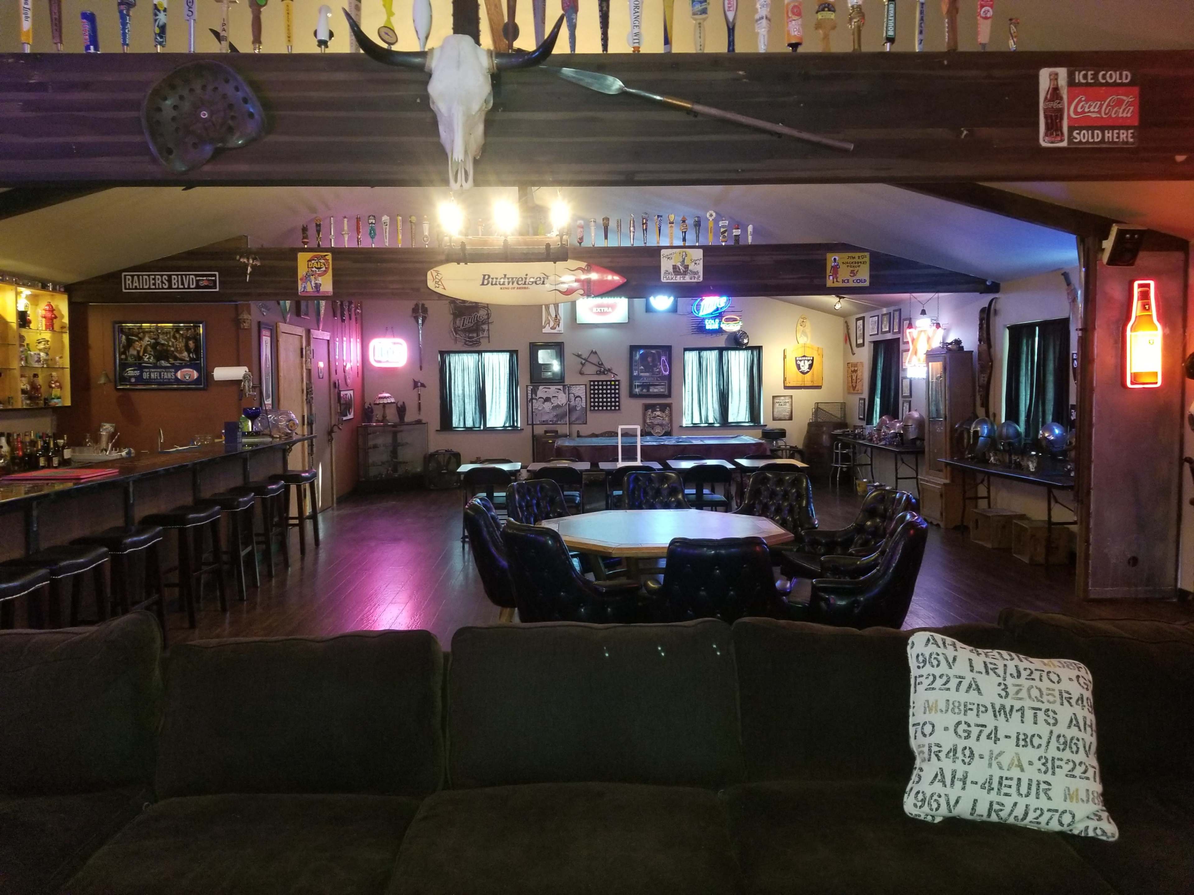 A spacious bar and gathering area with a central table surrounded by chairs, illuminated by neon signs and various decorations on the walls.