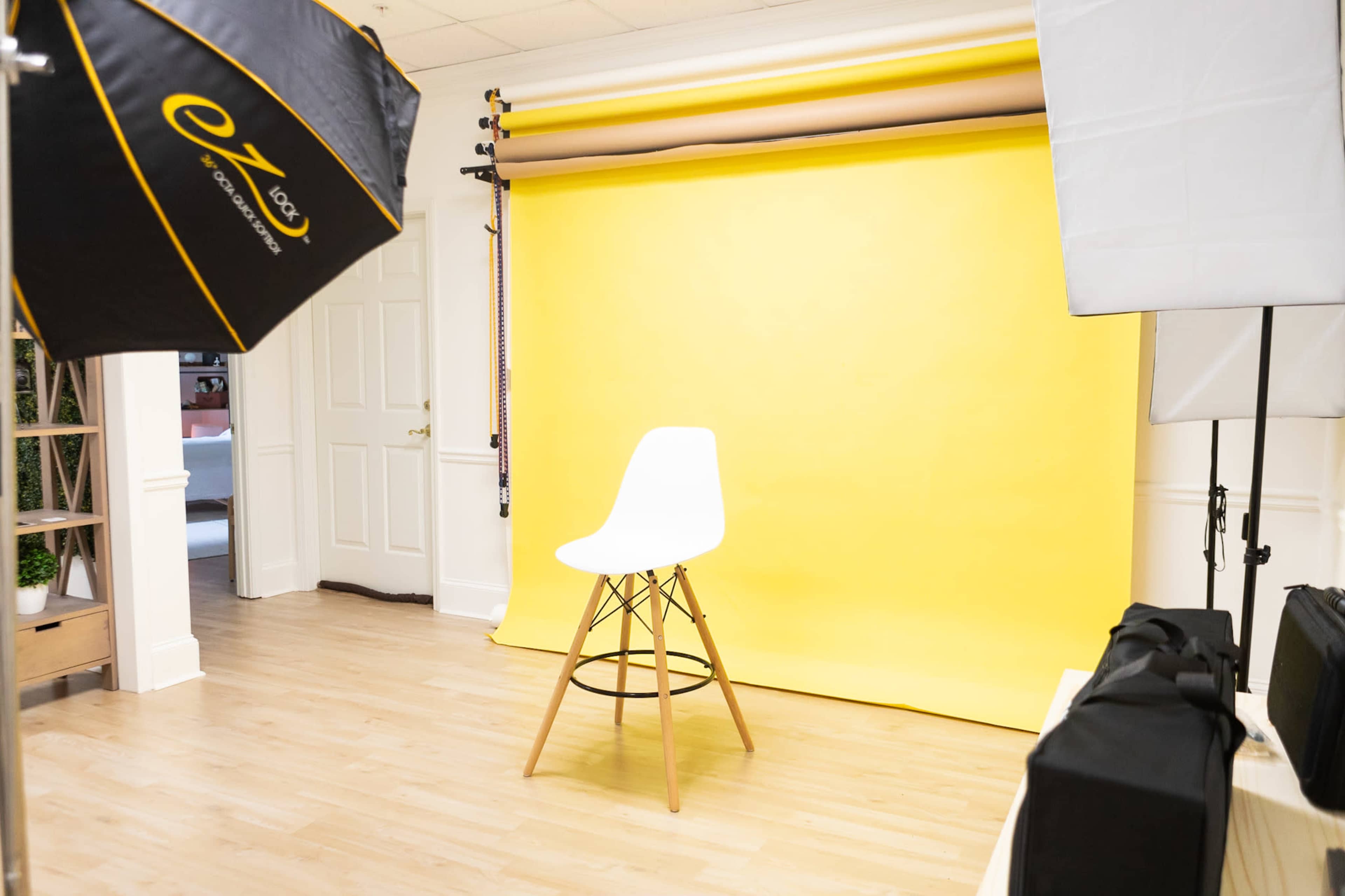 The image shows a photography studio with a white chair in front of a bright yellow backdrop, surrounded by lighting equipment and a neutral floor.