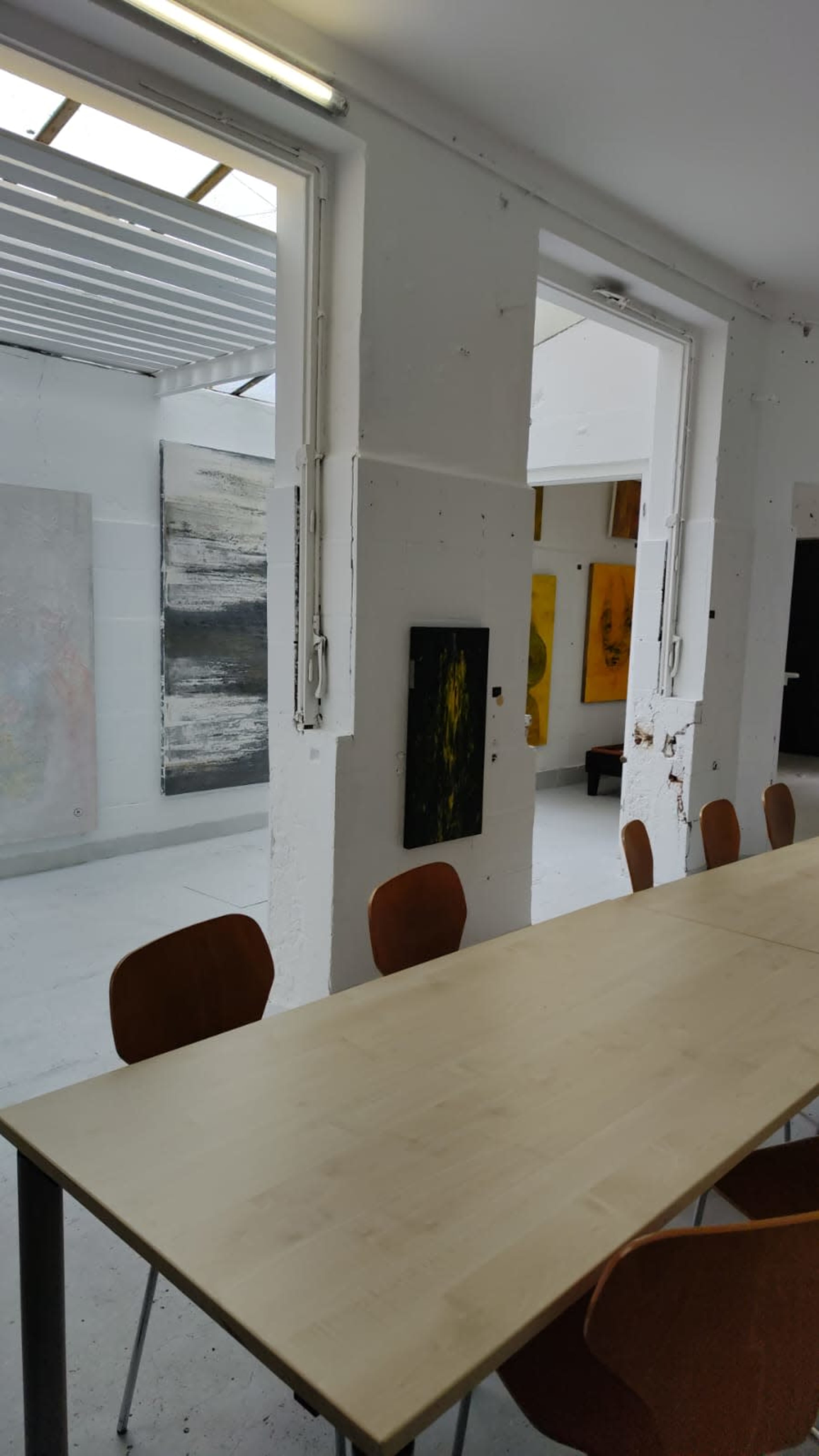 A spacious art gallery features a long wooden table surrounded by chairs, with various paintings displayed on the walls.