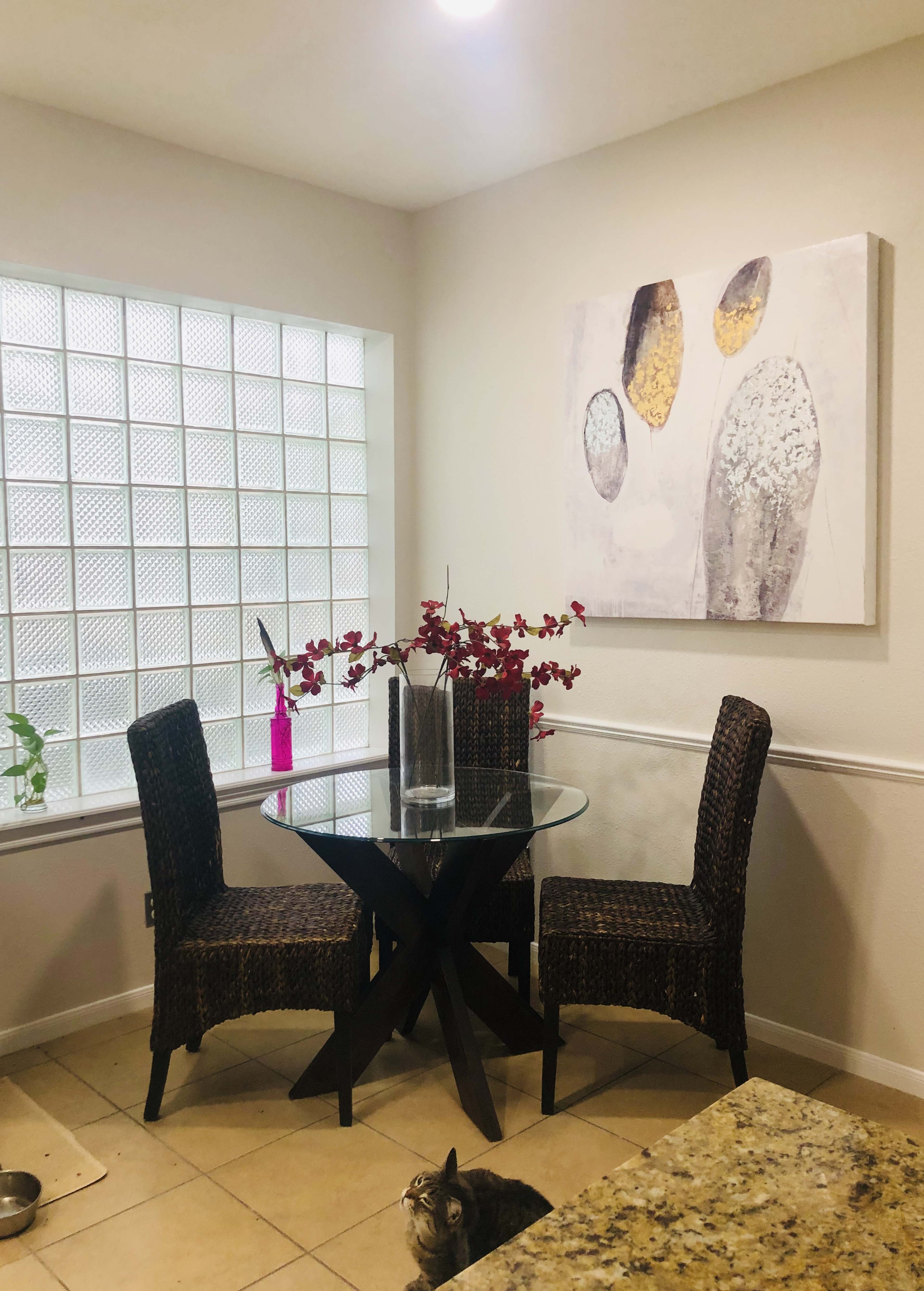 A small dining area features a round glass table with two woven chairs, surrounded by a plant and a vase of red flowers, with a cat resting on the tiled floor.