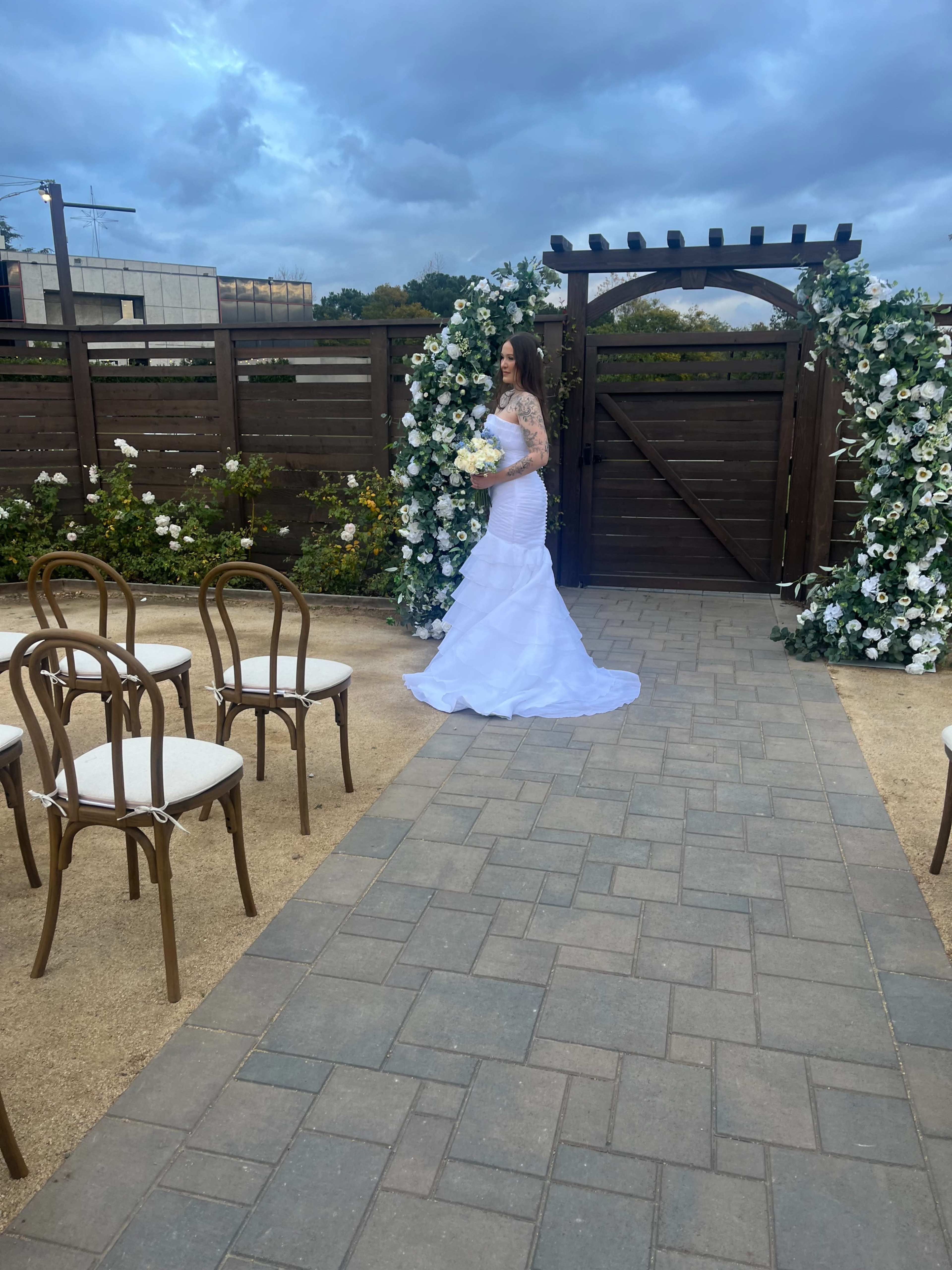 A bride in a white gown stands near a floral archway in an outdoor setting with wooden fencing and empty chairs arranged on a pathway.