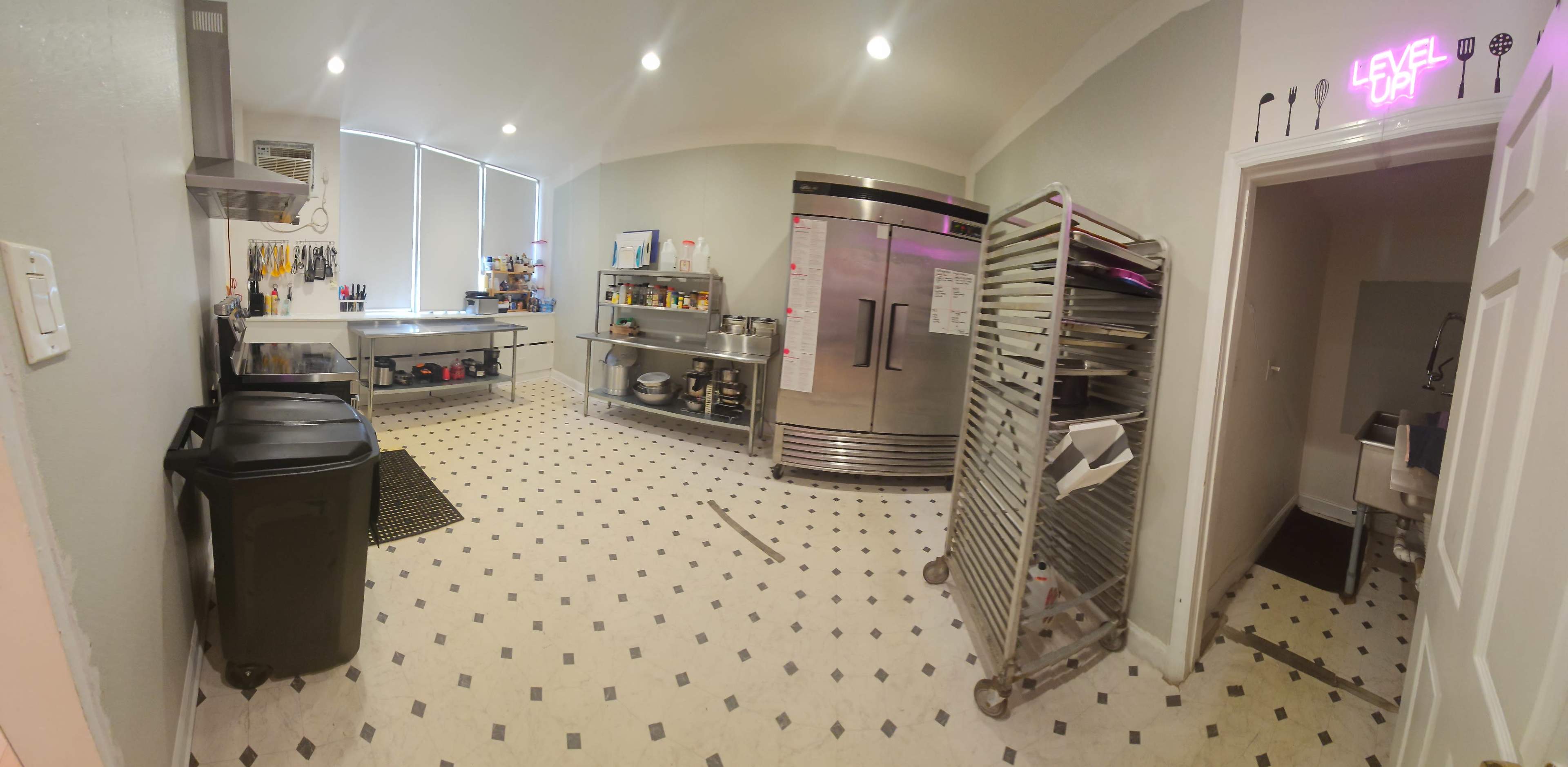 A spacious commercial kitchen with stainless steel appliances, worktables, and a storage area featuring racks and a doorway leading to another room.