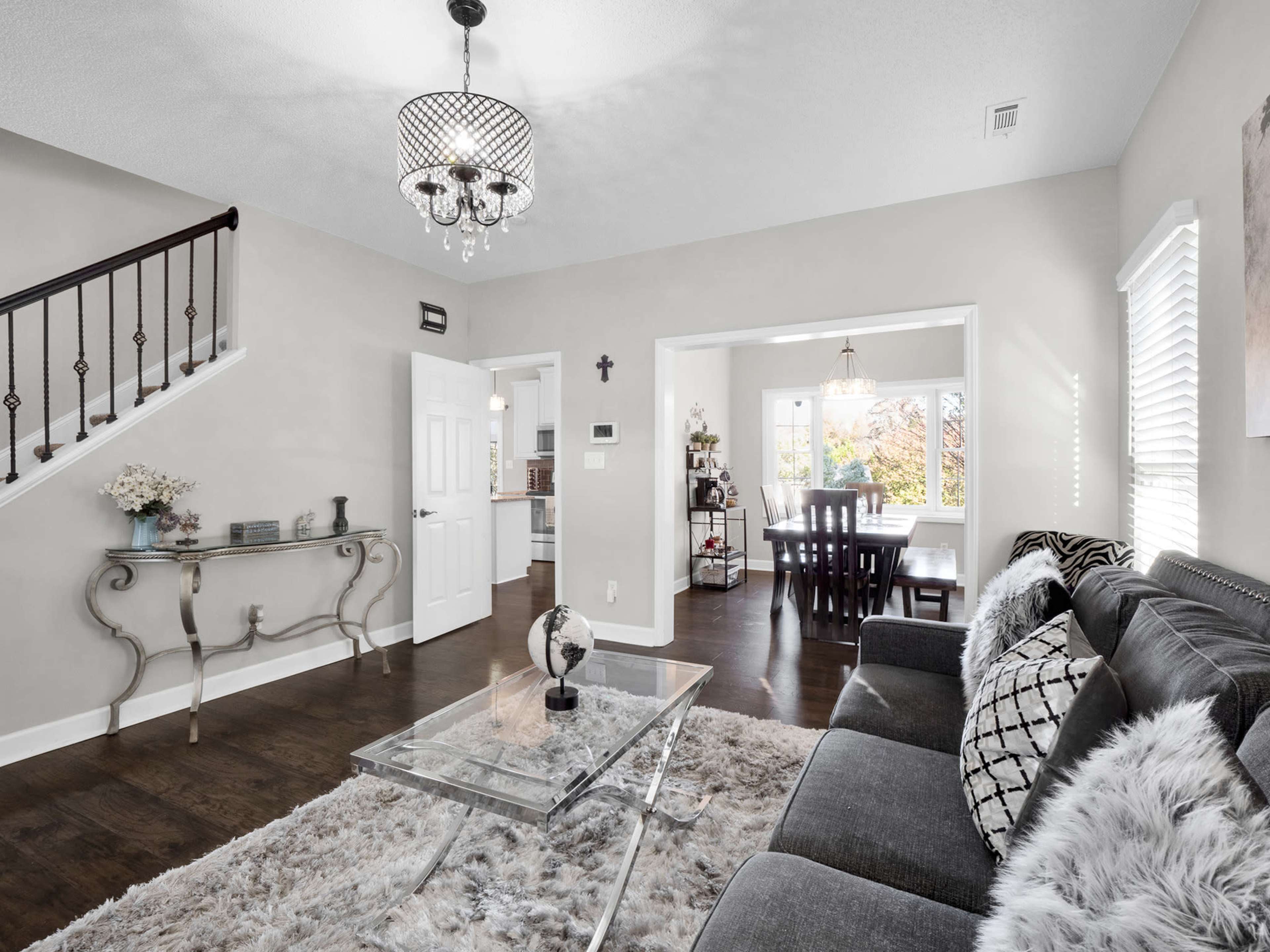 A well-decorated living room with a gray sofa, a glass coffee table on a plush rug, and an adjoining dining area visible through an open doorway.