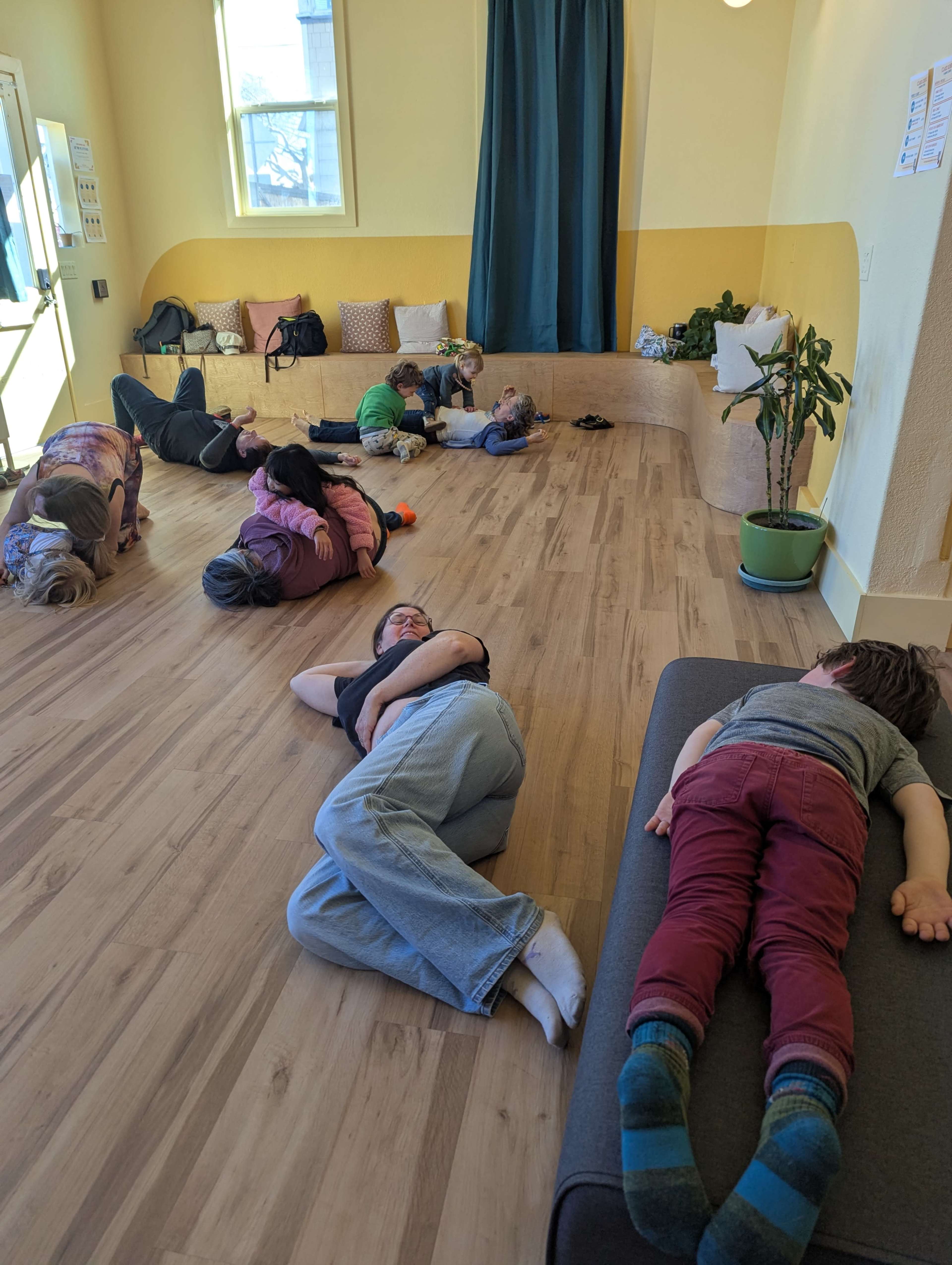 A group of children and adults are lying on the floor of a brightly colored room, with some resting on mats and others on the wooden floor.