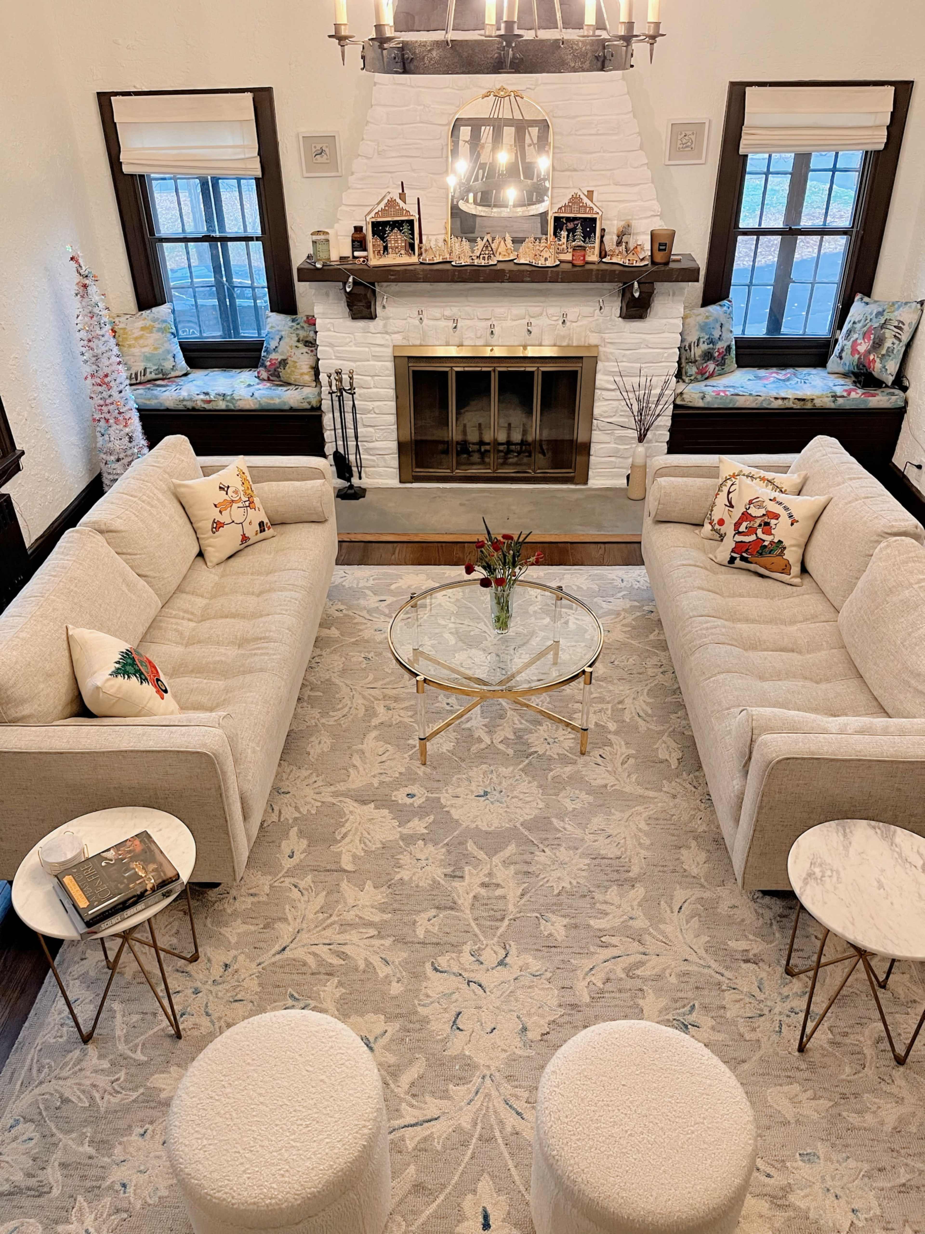The image depicts a cozy living room with two beige sofas facing each other, a round glass coffee table in the center, and a white-brick fireplace decorated with small houses and a Christmas tree.