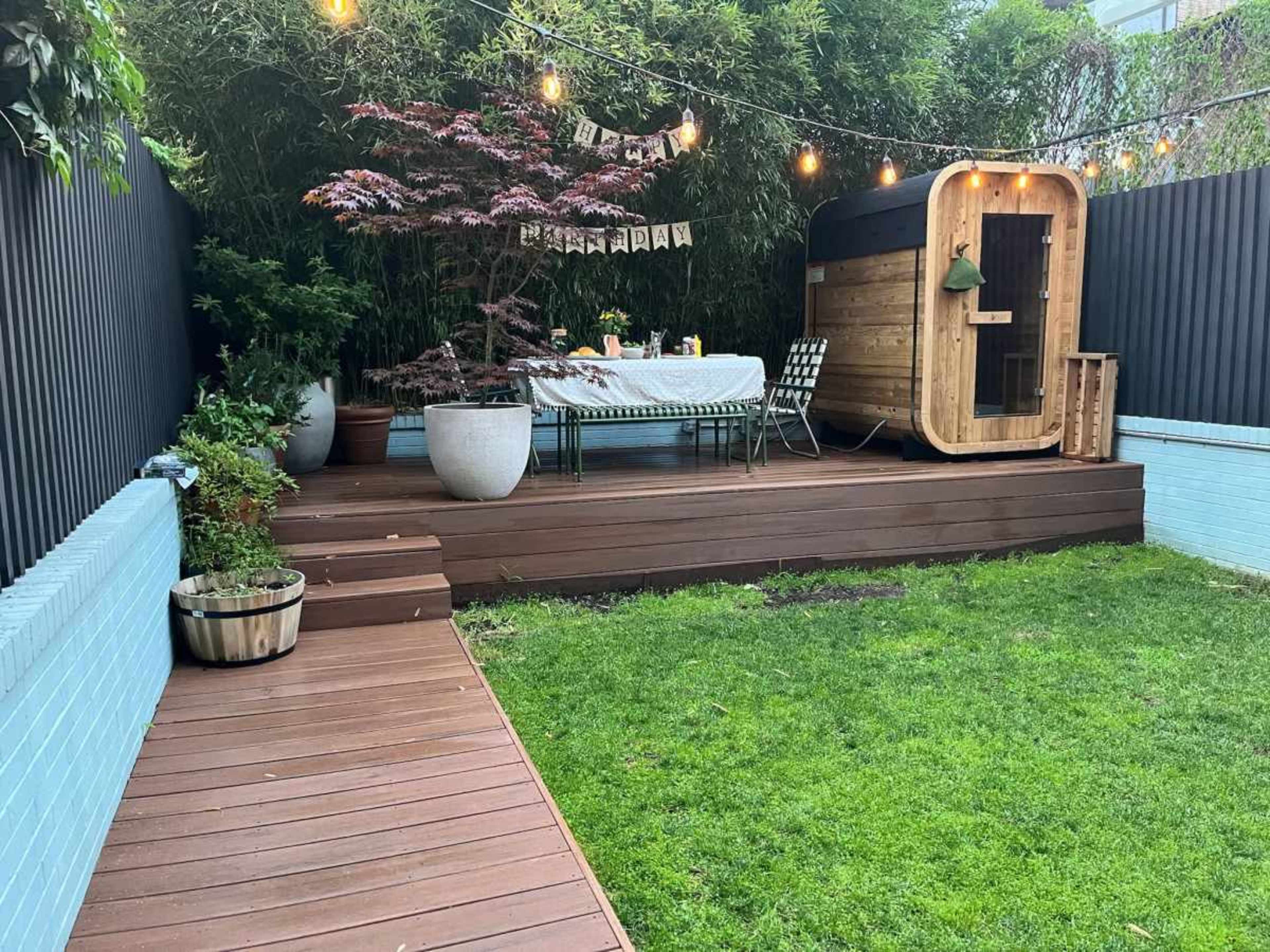 The image shows a backyard deck with a dining area, decorative string lights, potted plants, and a wooden structure resembling a sauna.