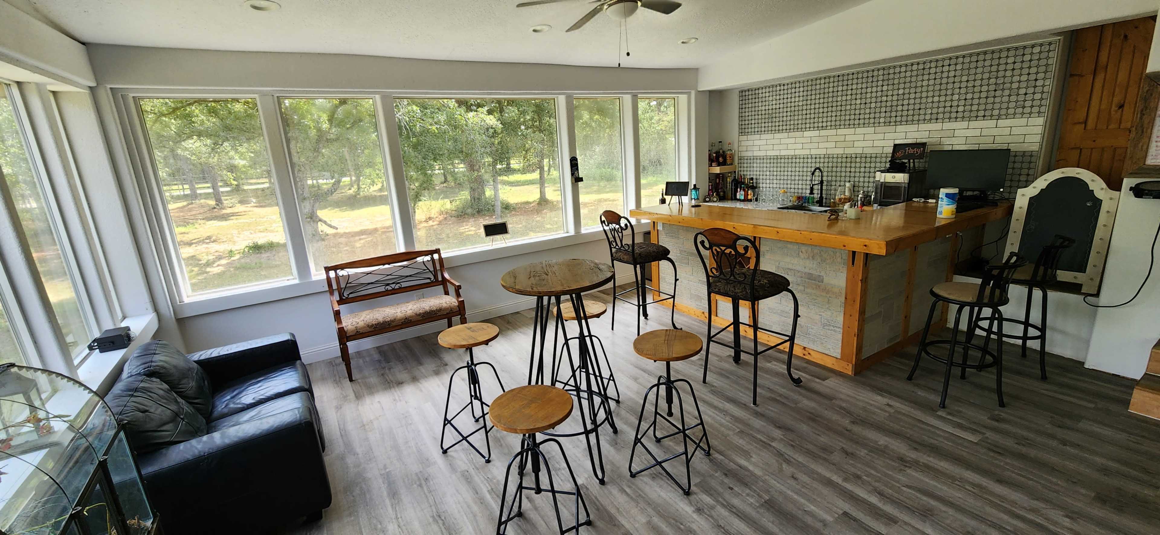 The image shows a bright, modern bar area with a wooden countertop, several metal bar stools, a sofa, and large windows allowing natural light and views of the outdoors.