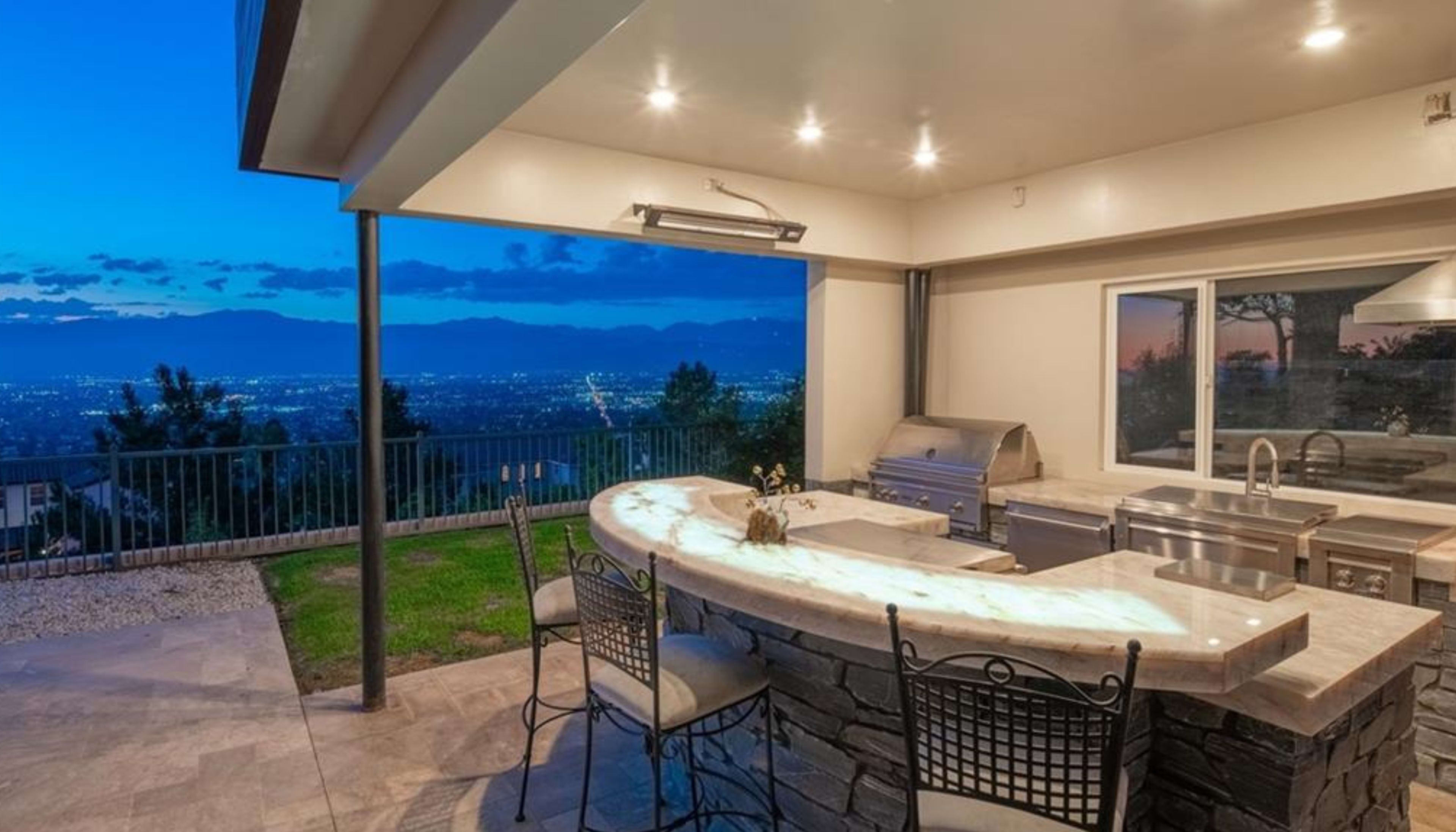 The image shows an outdoor kitchen with a bar area and a view of a city skyline at dusk.