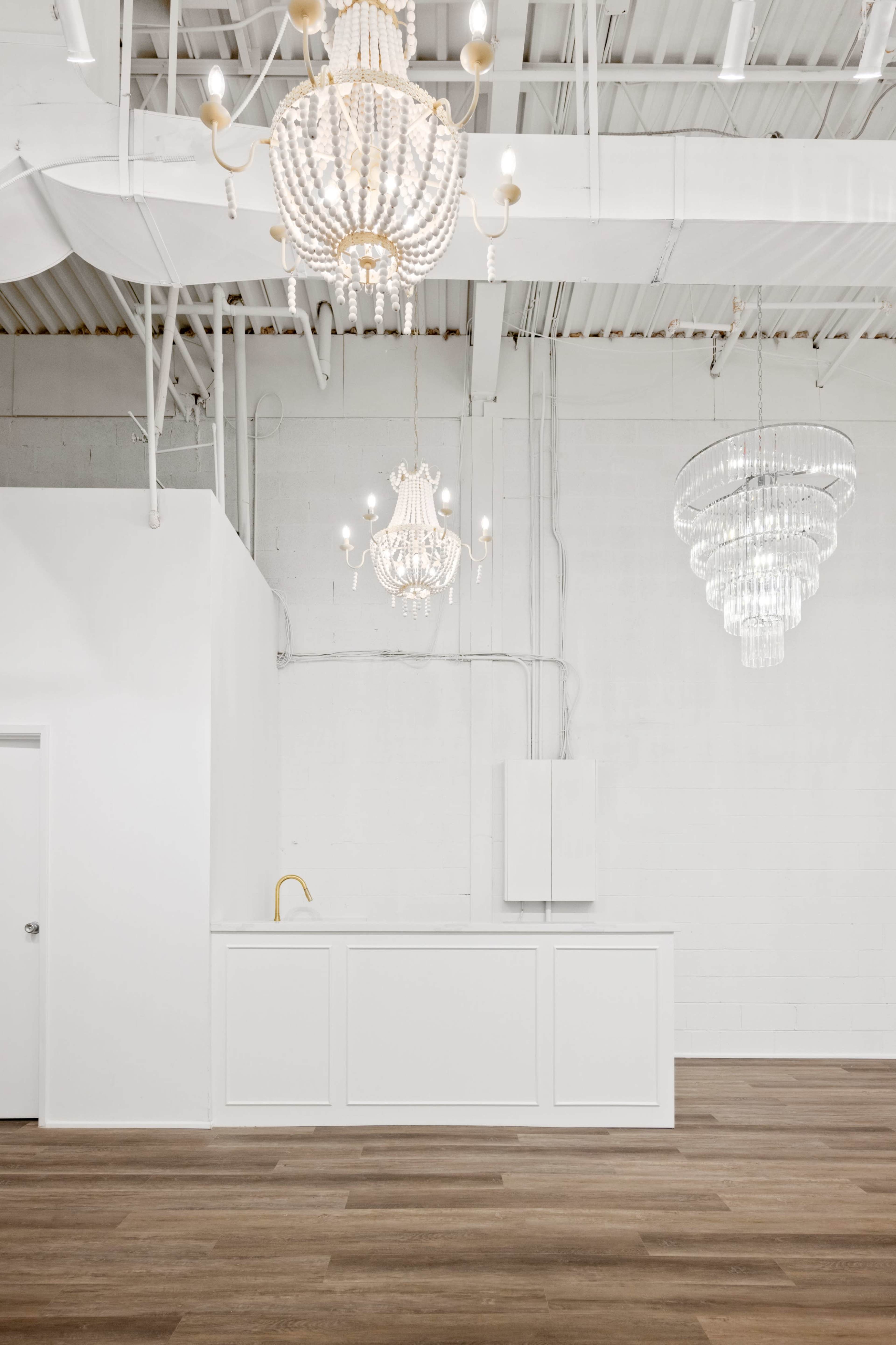 The image shows a spacious, well-lit room with white walls, wooden flooring, and multiple decorative chandeliers hanging from the ceiling.