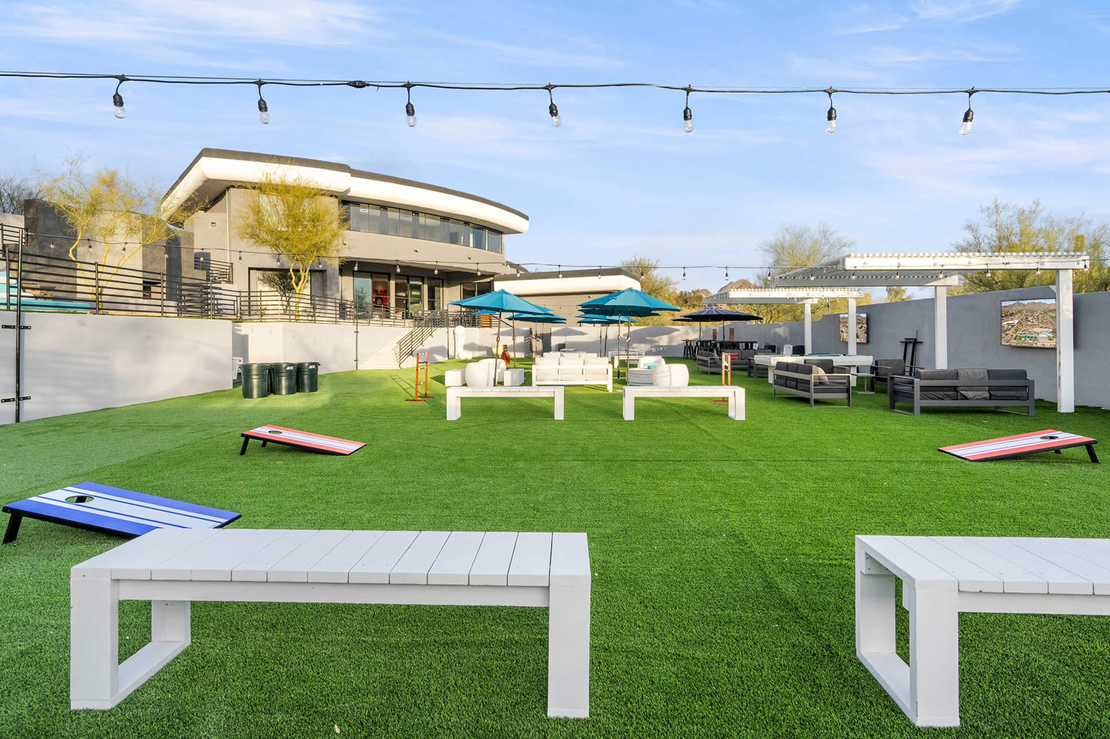 The image shows a landscaped outdoor area featuring artificial grass, several seating arrangements, and umbrellas, with a modern building in the background.